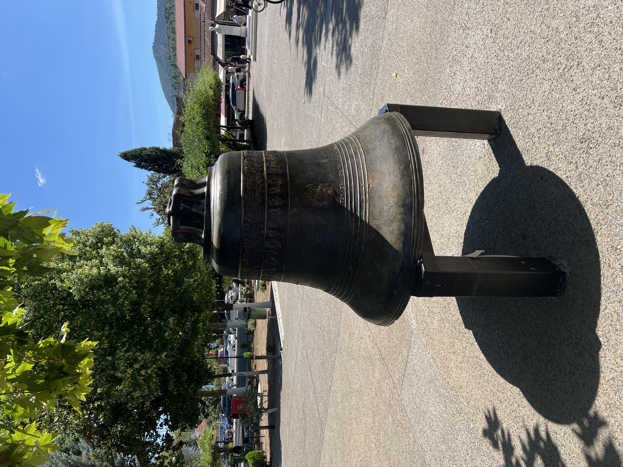 A large, dark bronze bell rests on a black metal stand in an open paved area. The bell is decorated with intricate patterns and lettering. The background shows a town square with trees, buildings, parked cars, and a few people. The sky is clear and blue. The bell is located in the town of Prokopi, Greece.