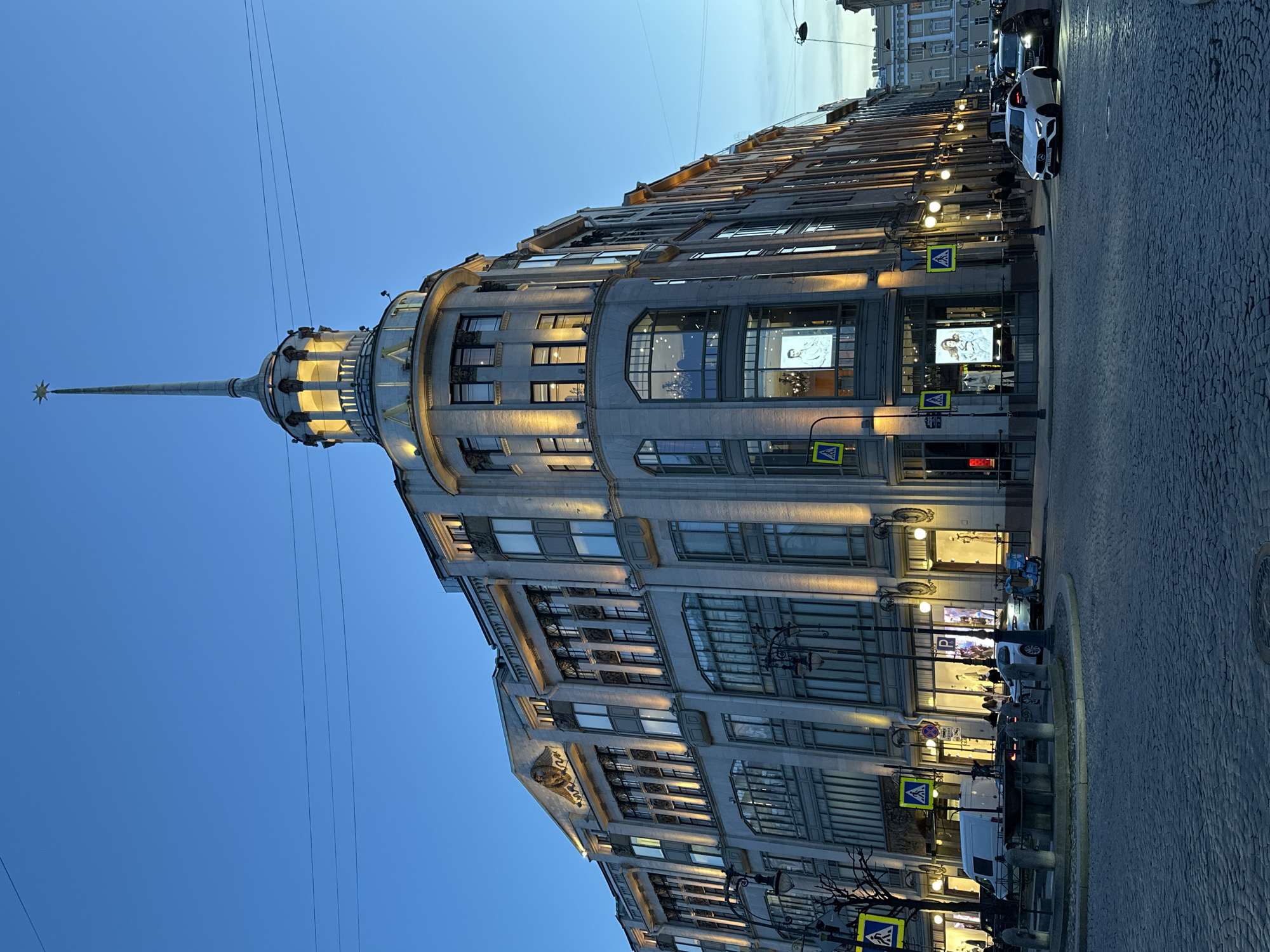 The image depicts the "Singer House," also known as the "House of Books," a prominent building located on Nevsky Prospekt in Saint Petersburg, Russia. The building has a distinctive Art Nouveau architectural style, characterized by its large windows, ornate detailing, and a tower topped with a glass globe.
The building is illuminated, with the lights visible through the windows. The sky is a twilight blue. The image is taken from a low angle, looking upwards, with the building appearing to stretch towards the top of the frame. In the foreground, a street with cars and pedestrians is visible. Streetlights are also present, contributing to the overall lighting of the scene.