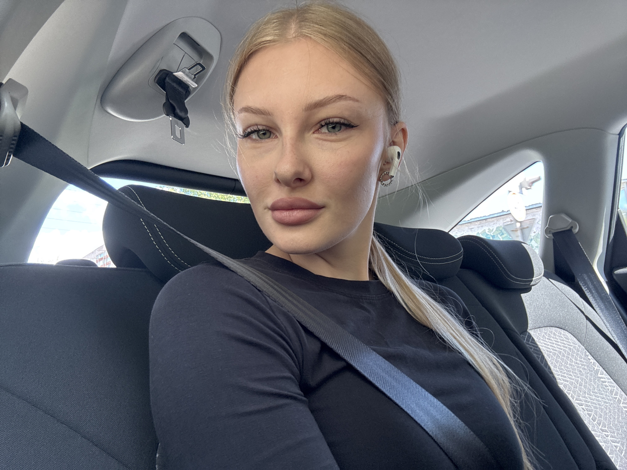 A blonde woman with green eyes and full lips is wearing a black long-sleeved shirt and a seatbelt inside a vehicle. She has on white wireless earbuds and is looking at the camera. The interior of the car shows black seats with white stitching and a grey headliner. Sunlight streams in from outside, illuminating the interior. The scene is set in a vehicle, likely in the city of Perm, Russia.