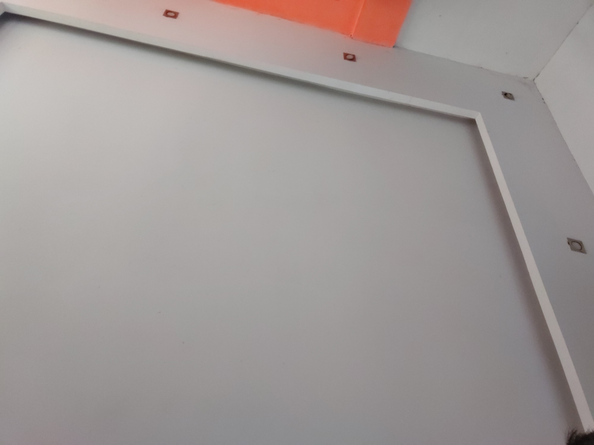 The image captures an upward-angled view of a modern interior wall and ceiling section. The primary subject is a large, rectangular surface painted in a uniform light grey, which occupies most of the frame. This grey surface is bordered by a clean white trim along its top and right edges, creating a framed effect.
Above the white trim, a vibrant orange or reddish-orange surface is visible, likely part of an adjacent wall or ceiling structure, providing a distinct color contrast. Along both the top white trim and the right white trim, several small, square-shaped indentations with darker centers are visible. These appear to be either recessed lighting fixtures or attachment points for architectural elements. Another similar indentation is located in the top-right corner where the ceiling meets the wall.
The setting is an indoor space, characterized by clean lines and a minimalist aesthetic. The surfaces appear smooth and recently finished, suggesting a contemporary room, possibly under construction or newly renovated. The lighting is even, indicating either soft natural light or internal artificial illumination. No specific activity or event is taking place, and no people are clearly visible, although a blurry, dark shape, possibly a person's hair or clothing, is present at the very bottom right edge of the image. There is no discernible text. The image provides no specific visual cues to indicate the location as Santa Ana, Venezuela.
