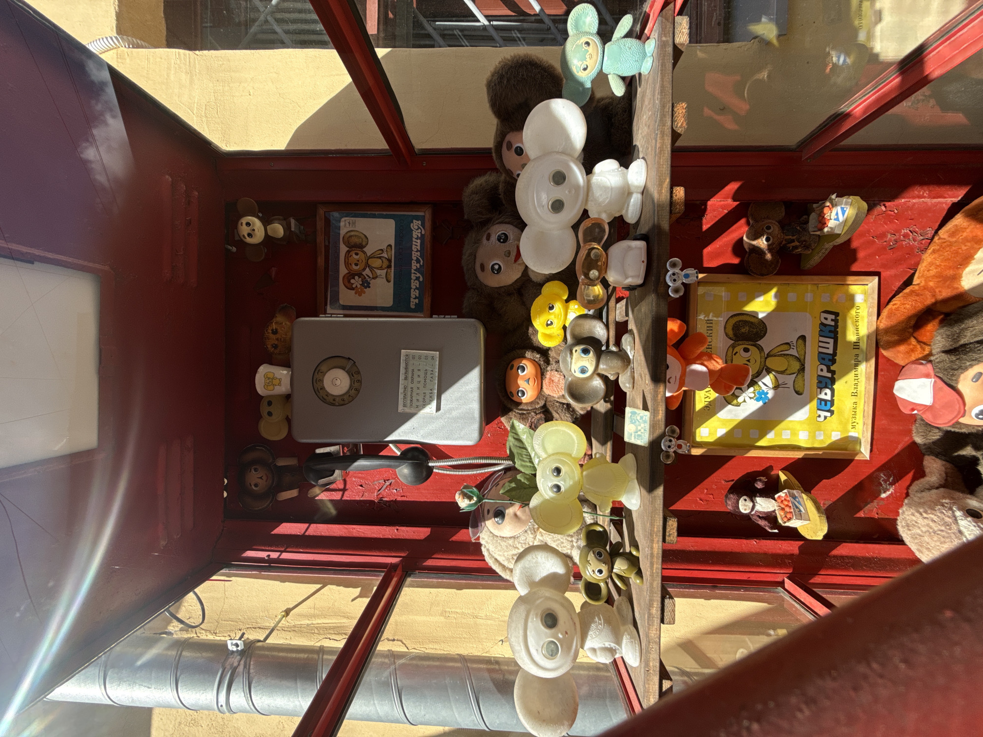 In Nizhny Novgorod, Russia, a red-painted telephone booth displays a collection of Cheburashka and Gena the Crocodile toys. The booth is set against a yellow wall, with a metal pipe running up the left side and a metal grate visible through the right side of the booth. The toys are arranged on shelves within the booth. On the top shelf, several small, lighter-colored Cheburashka-like figures and one brown monkey-like figure are visible, along with a vintage telephone receiver and a wall-mounted phone with a dial. The middle shelf holds a variety of Cheburashka figures in different sizes and colors, including large white and yellow plastic figures, smaller brown and green figures, and several fuzzy, brown, monkey-like plush toys. A framed picture of Cheburashka is also displayed on this shelf. The bottom shelf contains more plush toys, including a large orange and brown Cheburashka, a small figure of Cheburashka on a yellow boat with red fruit, and the lower parts of several other plush toys are visible at the very bottom of the frame. The scene is illuminated by sunlight, casting shadows and creating lens flare effects.