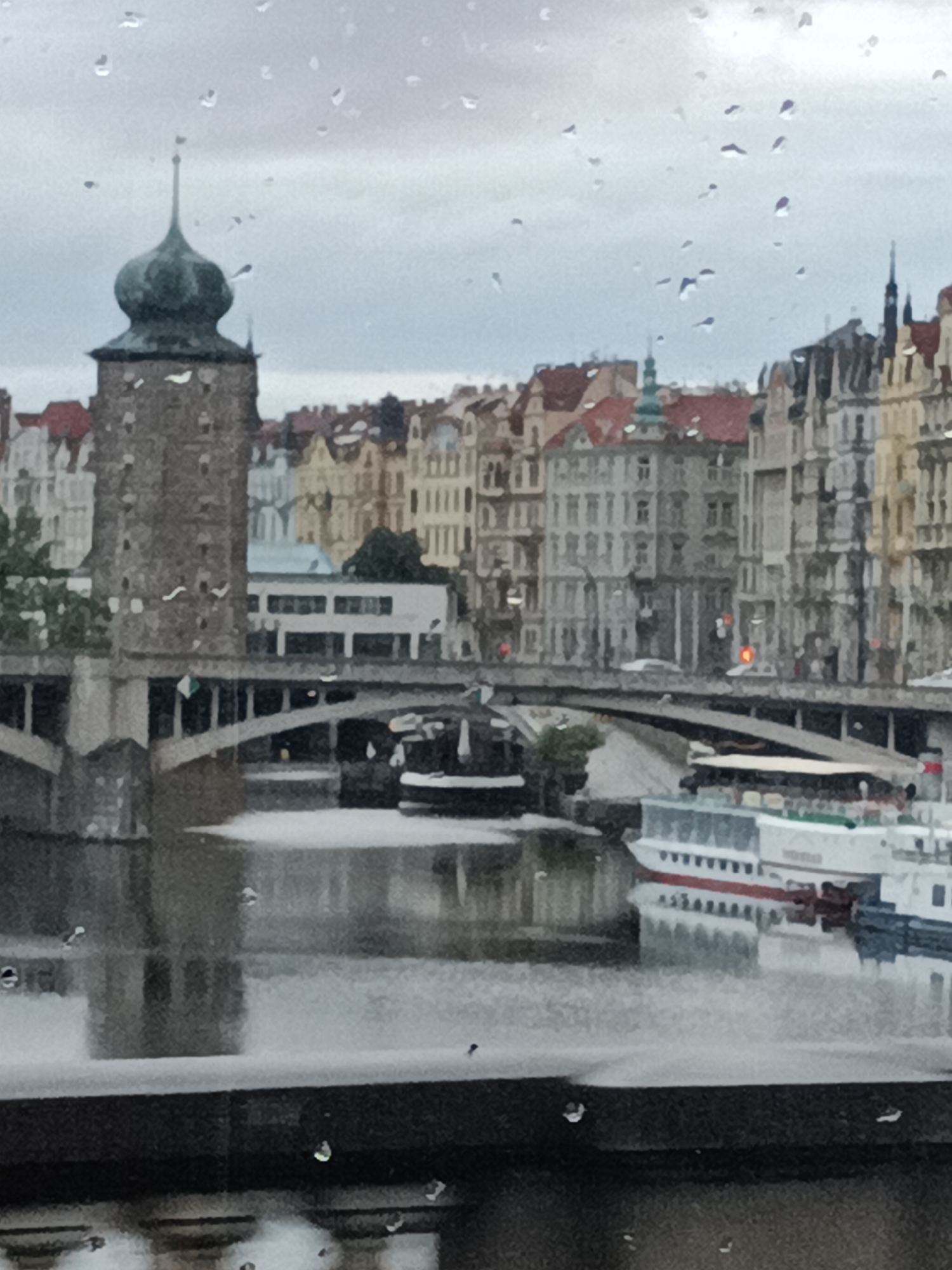 The image depicts a view of the Vltava River in Prague, Czechia, viewed through a window with raindrops. In the foreground, a dark, textured structure, possibly a ledge or balcony, is visible, with several raindrops on its surface. The river flows in the midground, with a white passenger boat docked on the right side. A bridge spans the river, with a tall, dark tower on the left side, topped with a green dome-like structure. Beyond the bridge and tower, a line of colorful, historic buildings with red roofs stretches across the cityscape. The sky is overcast and grey, with more raindrops visible on the windowpane.