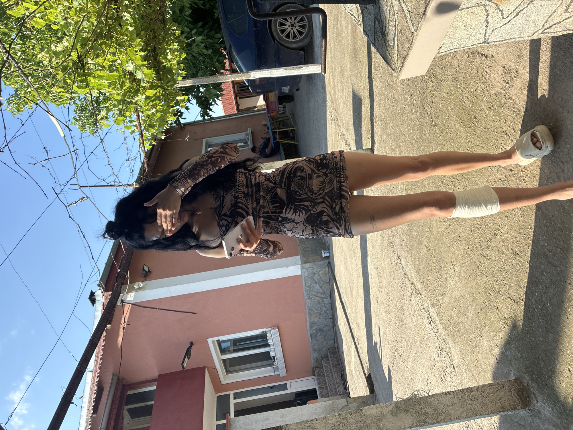 A young woman stands outdoors in Radievo, Bulgaria, on a clear, sunny day. She has dark hair styled partially in a bun and wears a short, form-fitting dress with a brown and black pattern resembling body art sleeves. Her right hand covers her face, while her left hand holds a silver smartphone with an illuminated screen. A white bandage is wrapped around her left calf, and she wears a light-colored slide sandal on her right foot, with her left foot bare. Visible tattoos are present on her left thigh.
The ground is concrete or asphalt, exhibiting cracks and debris, strongly illuminated by sunlight, casting distinct shadows. In the mid-ground, a salmon-colored building with white trim around windows and a door is visible, featuring stone cladding at its base. An outdoor light fixture is mounted on this building. In the background, a man in a dark shirt and light shorts sits on a blue bench at a small table, facing the building. A dark blue sedan is parked on the right, with a small utility trailer attached or nearby. Overhead, a bright blue sky is visible through a network of power lines, bare branches, and green foliage from a tree or vine. Concrete posts are present in the foreground and mid-ground.