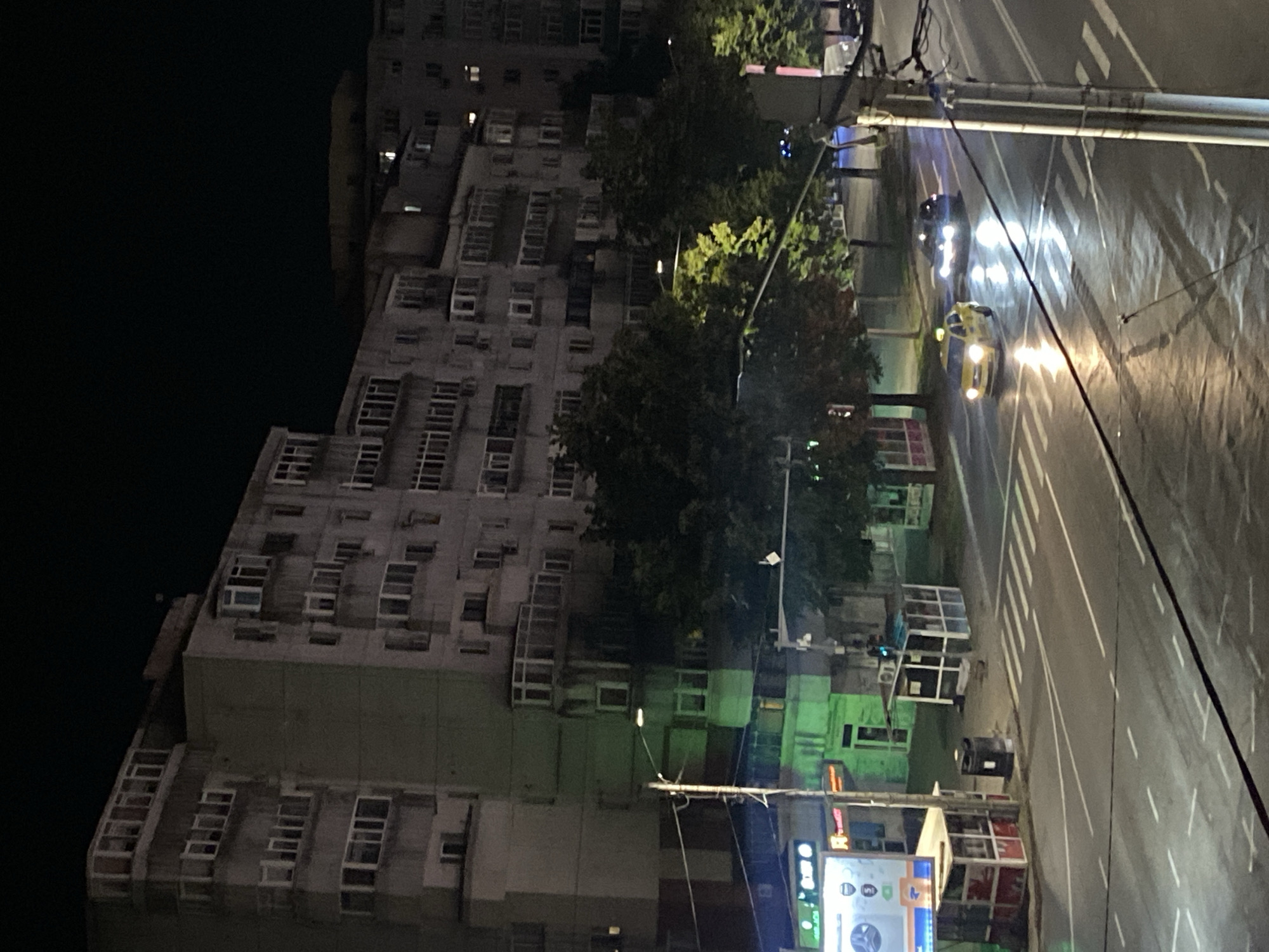 The image depicts a nighttime scene on a wet street in Iasi, Romania, likely near a residential area given the imposing apartment buildings. A yellow taxi is visible driving on the road, its headlights illuminated. In the background, several multi-story apartment buildings with numerous windows line the street. Some windows are lit, suggesting occupancy. Trees are present along the roadside, their leaves catching the ambient light. A pedestrian crossing is marked on the wet asphalt, reflecting the streetlights. There are also some small kiosks or shelters on the sidewalk. The overall impression is of a typical urban environment at night, with the wetness on the road indicating recent or ongoing precipitation.
