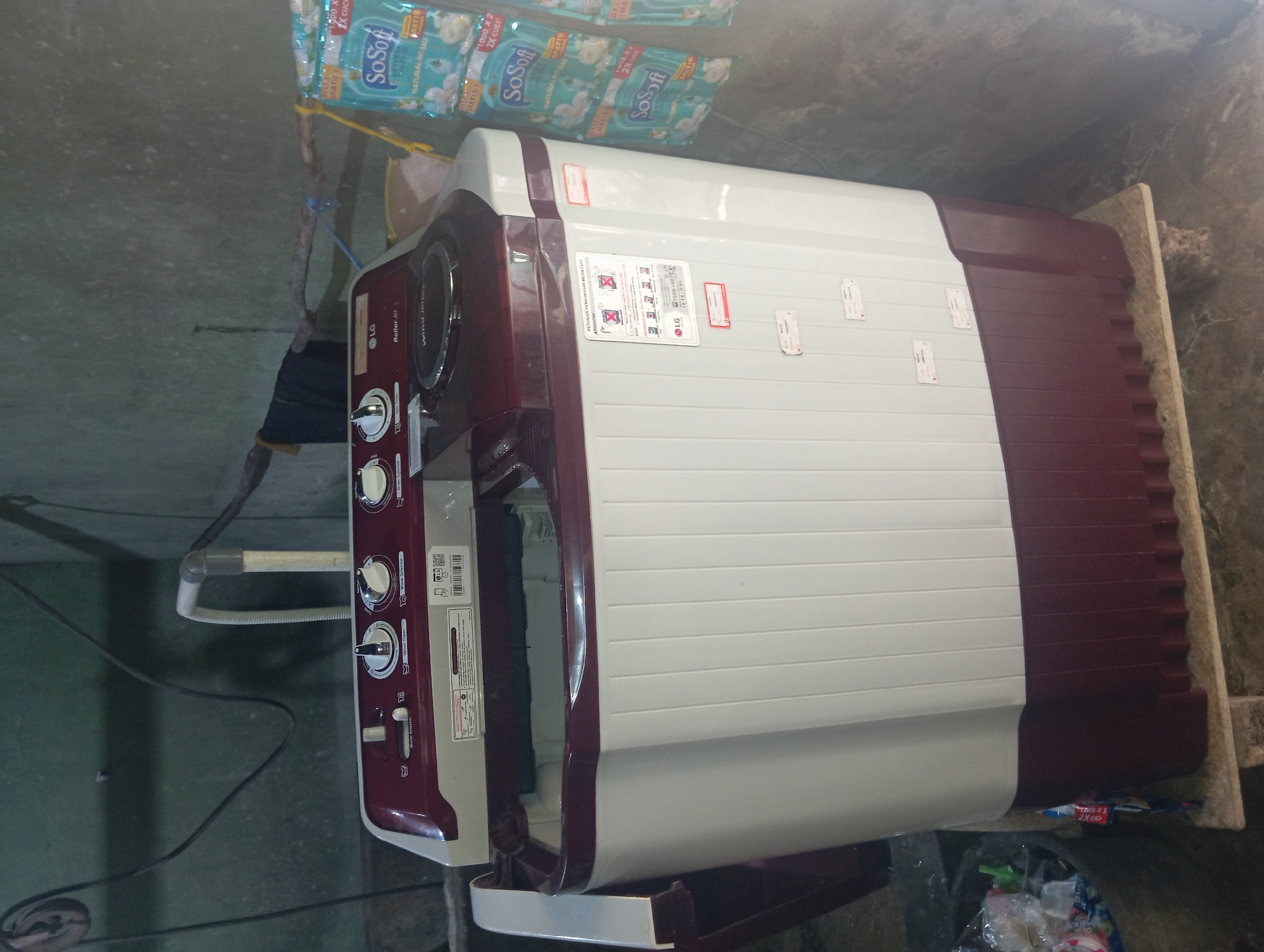 The image shows a LG semi-automatic washing machine in a dimly lit, cluttered indoor space, likely a utility area or backyard. The washing machine is primarily white with a maroon top and base. It has several control knobs on the top panel, labeled "Water Selector," "Wash Timer," and "Wash Selector." A circular spinner is visible in the center of the top. Several product labels are affixed to the washing machine, including one in Indonesian with diagrams. To the right of the washing machine, several packets of "So Soft" fabric softener are hanging, suggesting laundry is a recent or ongoing activity. A black garment is draped over a wooden stick supported by a pipe in the background. The floor appears to be concrete, and the walls are rough and unpainted. The scene suggests a functional, but not pristine, domestic environment in Rejosari, Indonesia.