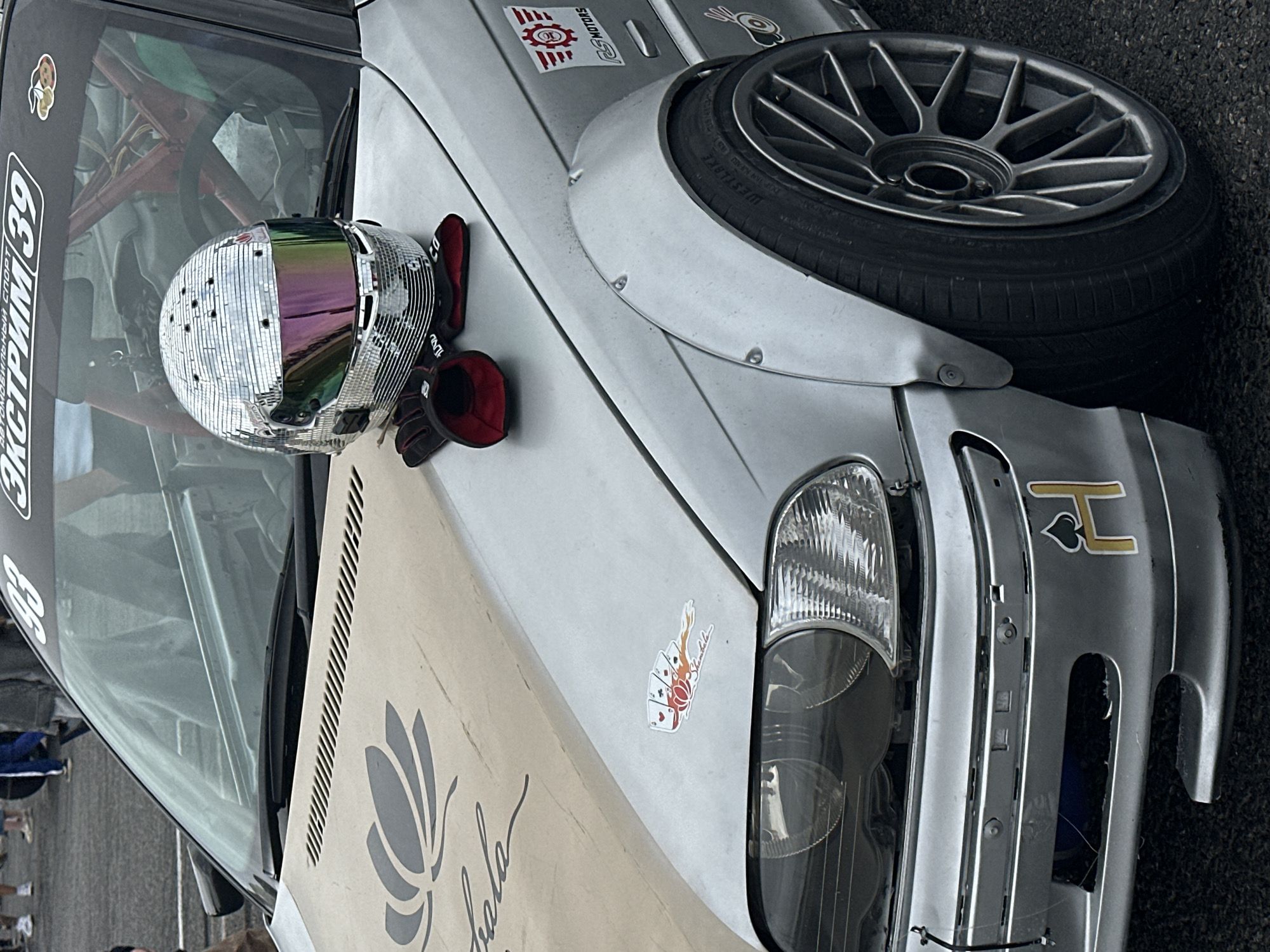 An exterior, eye-level, medium shot captures a modified silver racing car parked outdoors on asphalt, likely at an event. The car's hood displays a large, stylized grey lotus flower graphic and what appears to be a cursive word. Resting on the hood are a pair of red racing gloves and a silver, disco-ball-like helmet with a mirrored, iridescent visor. A racing cage is visible through the car's windshield. Stickers adorn the car, including a "RS MOTORS" logo on the fender and a playing card-themed decal on the hood near the headlight. The front bumper also features a sticker with the letter "H" inside a black spade. In the blurred background, several people are walking. The context suggests this scene is from Kaliningrad, Russia, given the presence of Russian text on a sticker at the top of the windshield, which reads "Автомобильный спорт 39", translating to "Automobile Sport 39", with "39" being a region code for Kaliningrad Oblast.