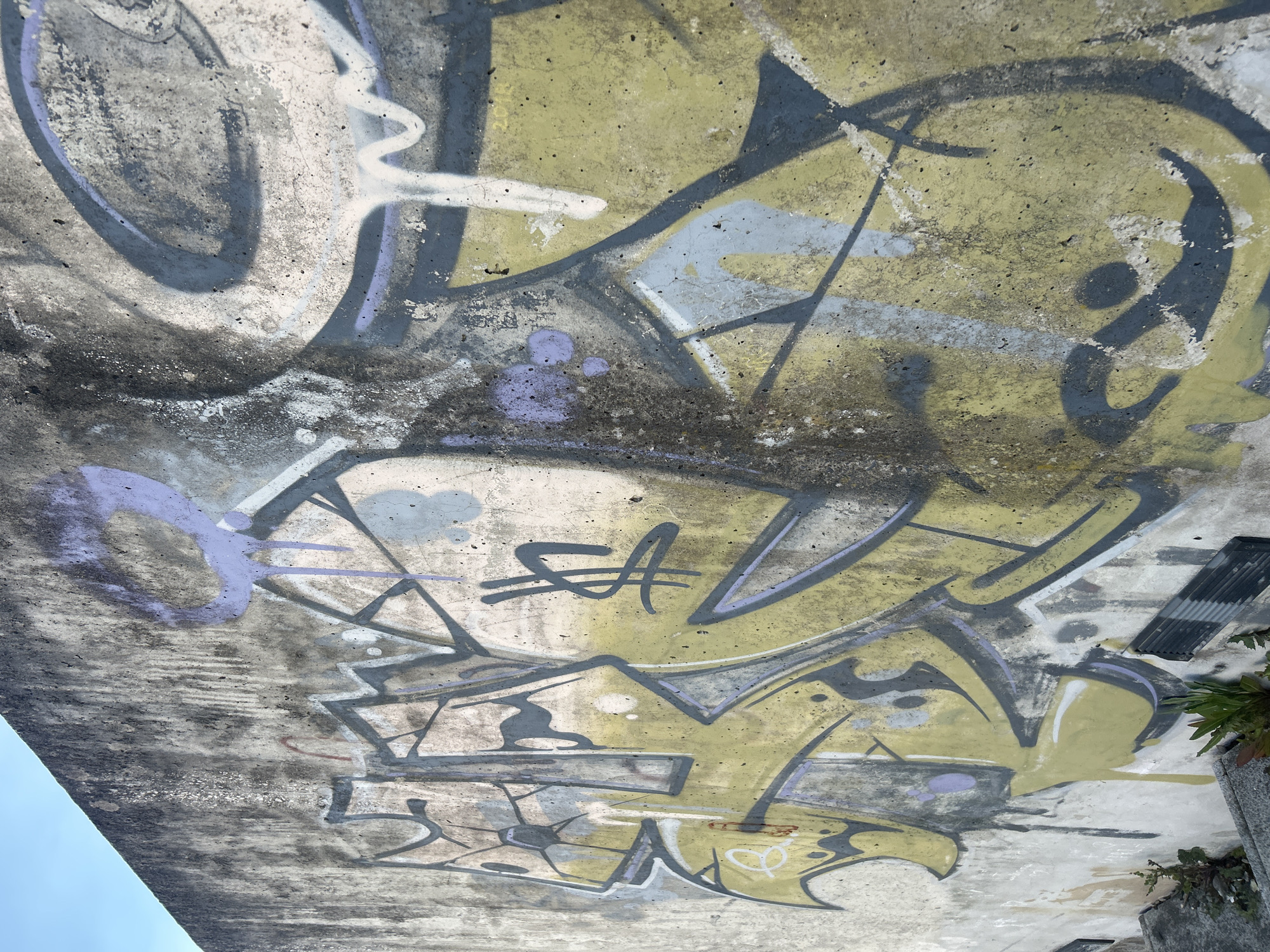 The image depicts a concrete surface covered in layers of graffiti. Predominant colors in the graffiti include yellow, grey, purple, and white. The style is abstract, with overlapping lines and shapes that make it difficult to discern specific words or images. The concrete itself appears weathered, with visible cracks and discoloration. Part of a blue sky is visible in the bottom left corner. In the upper right corner, there are some objects, possibly metal structures and some vegetation.
This type of graffiti is often found in urban areas, such as Vila Real, Portugal.
