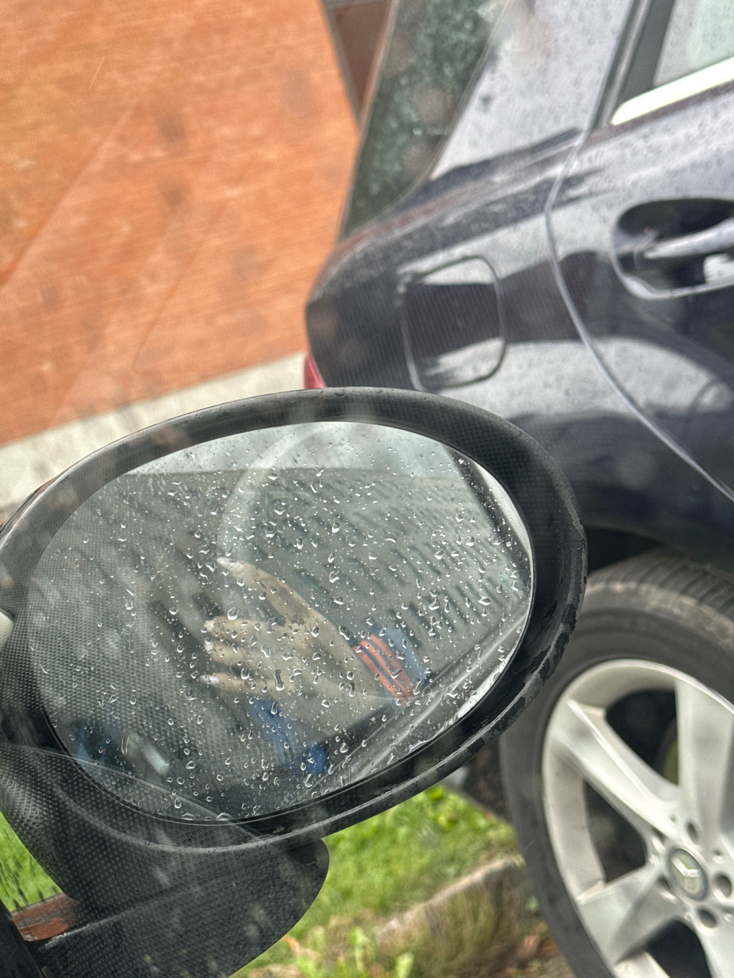 A close-up shot captures a car's side mirror, reflecting raindrops and a portion of the vehicle's dark exterior. The mirror's housing appears to be made of a textured black material, possibly carbon fiber, with some visible wear and tear. Behind the mirror, a dark-colored car, likely a sedan, is parked, with its fuel cap and door handle visible. The car's body is wet, indicating recent rain. A silver alloy wheel with a Mercedes-Benz logo is also in view. The background shows a blurred reddish-orange surface, possibly a wall or building, and some greenery, suggesting an outdoor urban environment in Tallinn, Estonia.