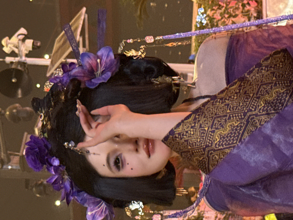 A woman with fair skin and dark hair adorned with purple flowers, delicate ornaments, and trailing strands poses elegantly. Her hand is raised near her face, partially obscuring her eye. She is dressed in a purple garment with gold geometric patterns on the sleeve. The background is softly blurred, suggesting an outdoor or event setting with lighting equipment visible.