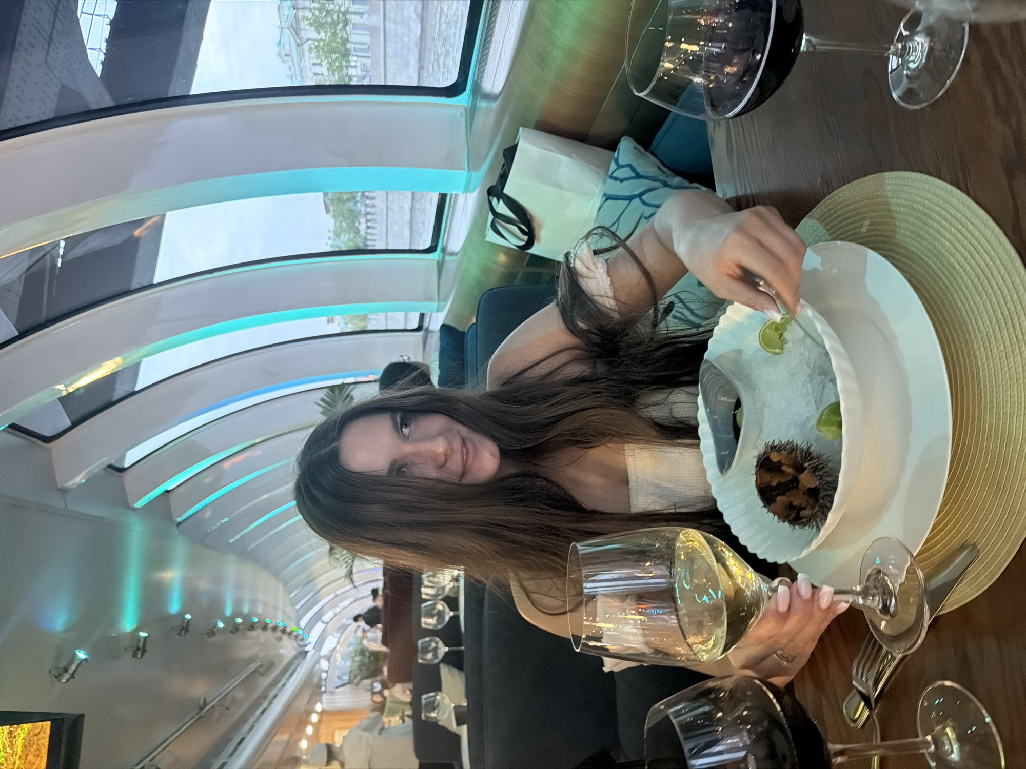 A woman with long, dark hair sits at a table on a boat, likely a river cruise in Moscow, Russia, given the architecture visible through the windows. She is wearing a white strapless top. In front of her is a white bowl containing what appears to be sea urchin on ice, with a slice of lime. She is holding a fork, delicately touching the lime. Two wine glasses are on the table: one with white wine in front of her, and another with red wine to her right. In the background, other diners are seated, and the interior of the boat features curved, modern lighting with blue and green hues. A white shopping bag rests on a blue cushion behind her. The river outside the window shows buildings and trees lining the bank, characteristic of Moscow.