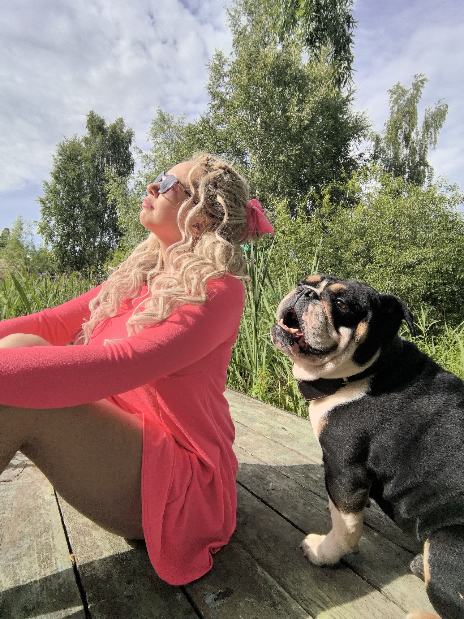 In a sunny outdoor setting, a woman with blonde, curly hair, wearing sunglasses and a bright pink dress, sits on a wooden platform. Her hair is styled with braids and adorned with a pink bow. She is looking upwards towards the sky. Beside her sits a black and tan bulldog, also looking up, with its mouth slightly open. The dog wears a dark collar. The background features lush green trees and tall reeds, suggesting a natural environment like a park or lakeside. The lighting indicates it is daytime, likely midday, with bright sunlight casting shadows on the wooden planks. The overall mood appears relaxed and peaceful, with both subjects seemingly enjoying the warm weather. There is no visible text in the image.