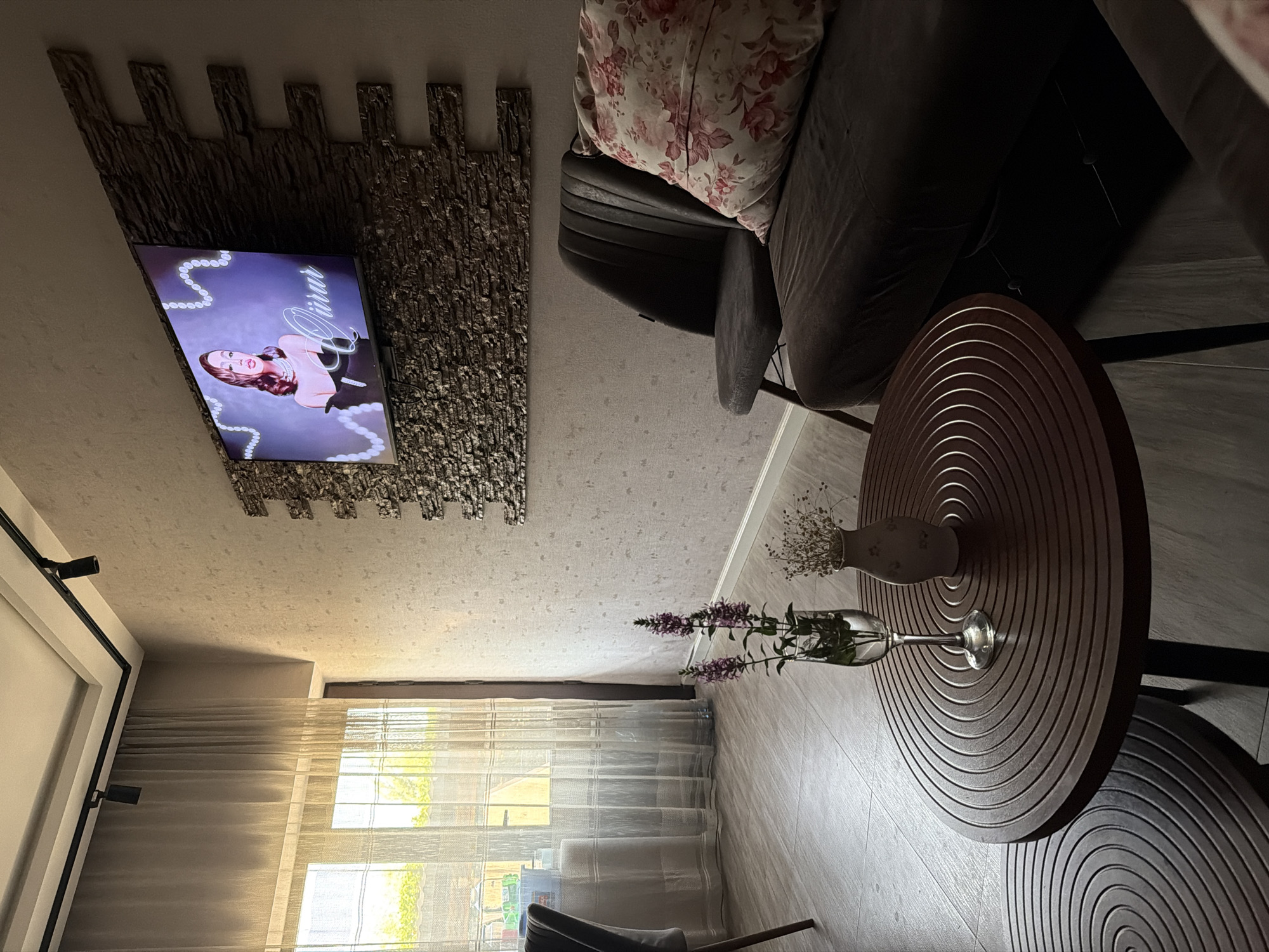 The image depicts an interior scene, likely a living room, with a focus on a television mounted on a textured wall above a sofa and a chair. The television displays a person in formal attire, possibly a singer or actress, with "Qürur" written in cursive below. A floral-patterned pillow rests on the dark-colored sofa. In the foreground, a round wooden coffee table holds two decorative items: a wine glass with purple flowers and a small vase with dried flowers. To the left, sheer curtains cover a window, allowing natural light to enter the room. The flooring appears to be tiled. The overall ambiance suggests a modern and somewhat artistic decor. The location is identified as Sabirabad, Azerbaijan. There are no individuals present in the scene.