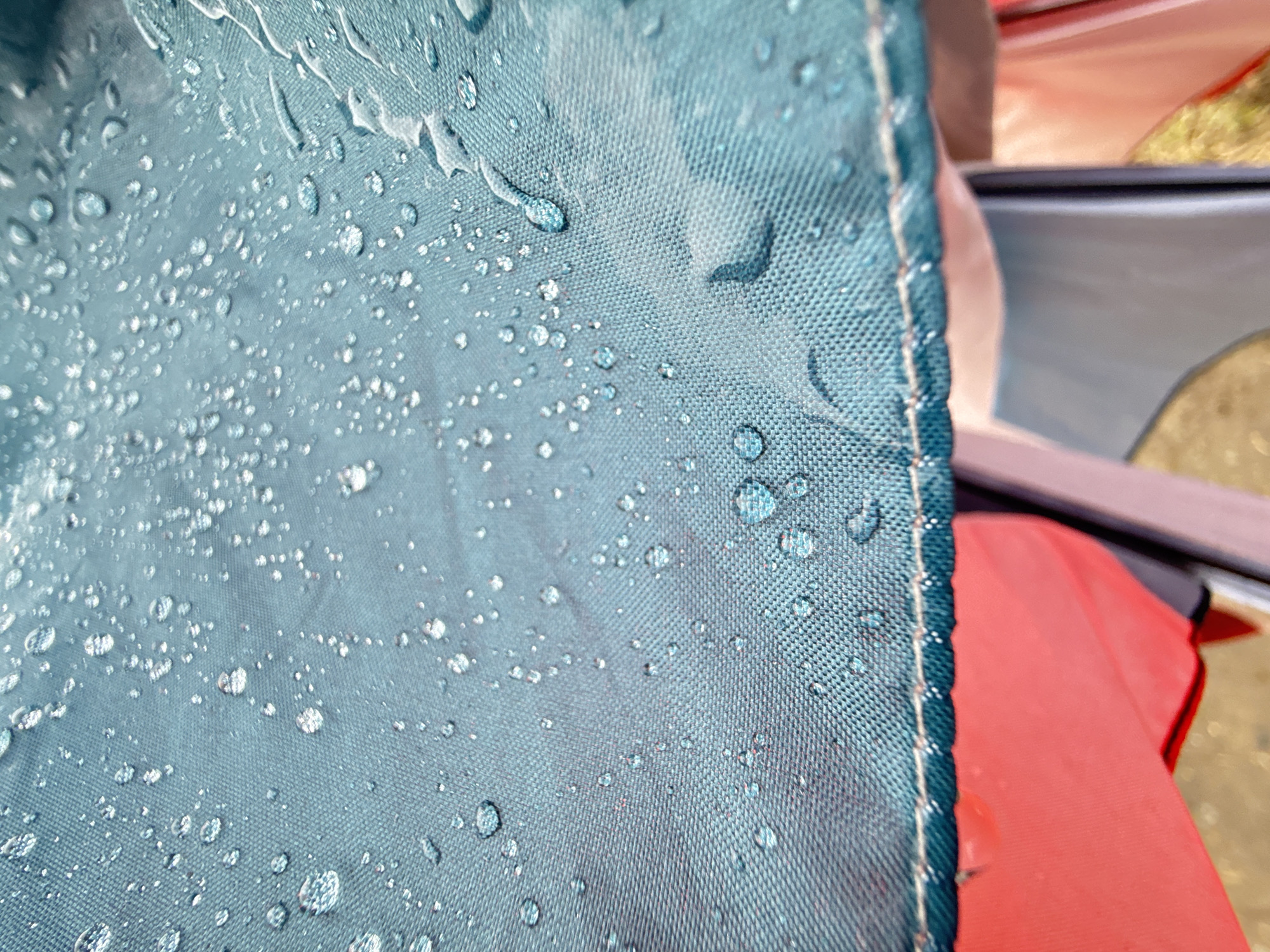 A close-up shot shows a blue-green waterproof tent fabric covered in water droplets. The droplets range in size, scattered across the textured surface of the fabric. A stitched seam runs along the edge of the fabric. Partially visible in the background is a red piece of fabric and other parts of the tent structure. The location is noted as the city of Dmitrov, Russia, but no information in the picture confirms the city.