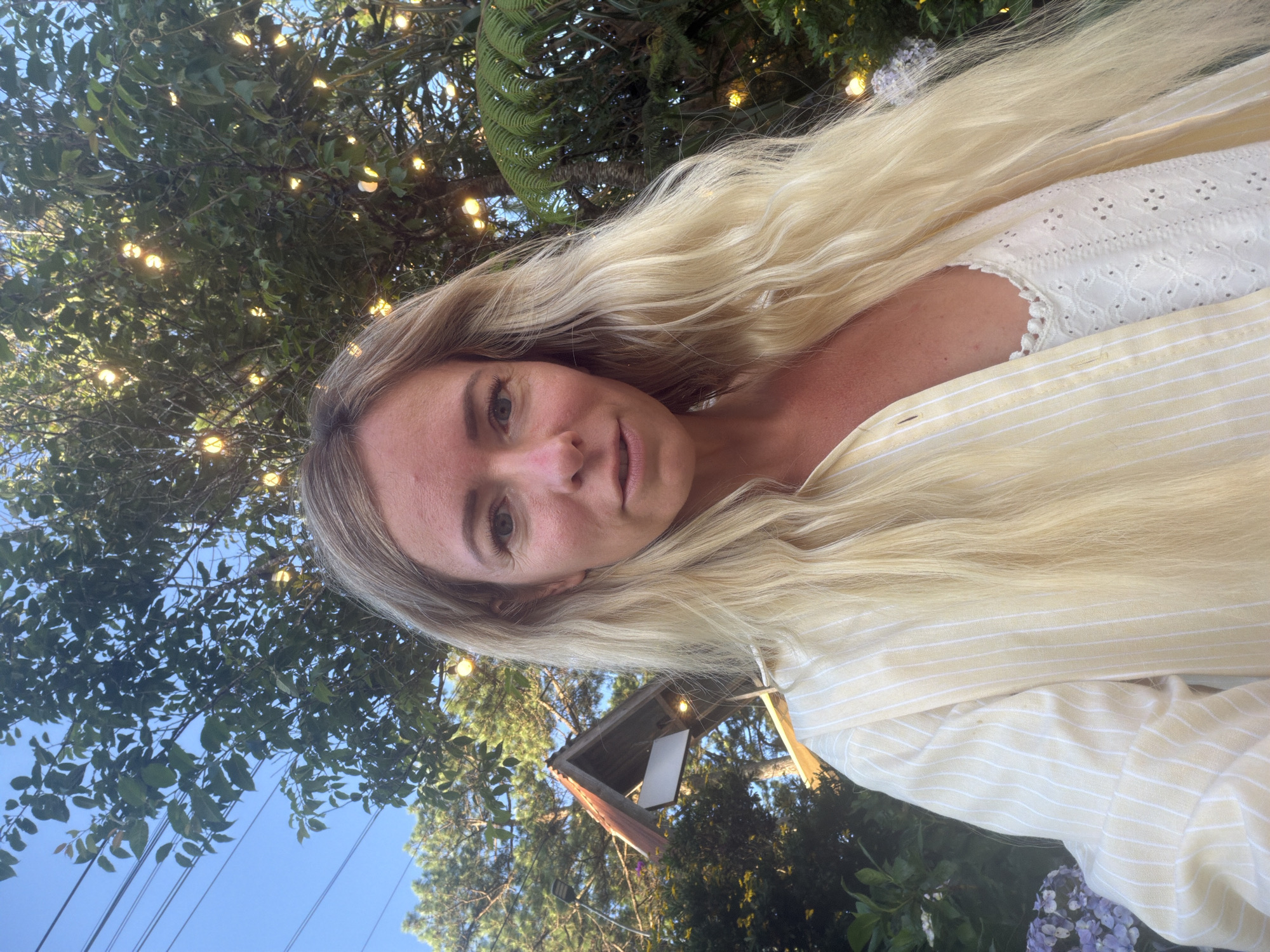 This is a selfie of a blonde woman taken outdoors in Xuân Thành, Vietnam. The woman is looking at the camera with a neutral expression. She has long, wavy blonde hair and is wearing a light yellow, vertically striped shirt over a white eyelet top. The background features lush green trees with fairy lights strung throughout them. A blue sky is visible above the trees, along with power lines and a small wooden structure with a slanted roof. There are also some purple flowers in the lower left of the frame. The lighting suggests it is either late afternoon or early evening.
