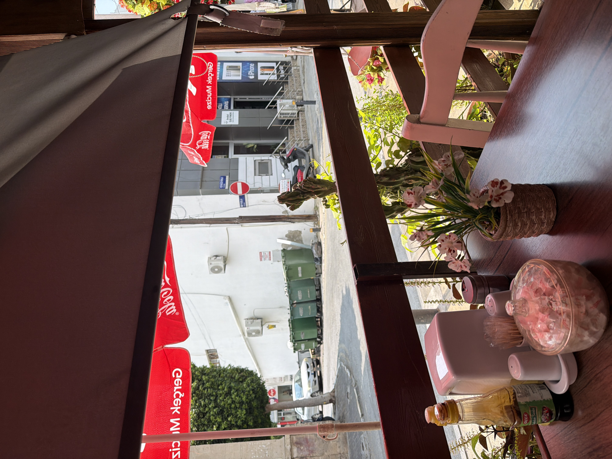 This is a photograph taken outdoors, likely at a cafe or restaurant in Kyrenia, Northern Cyprus. The image is oriented vertically, giving a sense of looking down from an elevated perspective. The foreground features a table with various items, including a bottle of what appears to be olive oil, a salt and pepper shaker set, a container of toothpicks, and a clear, decorative jar filled with pinkish items, possibly candy or potpourri. A pink chair is visible in the mid-ground, and a potted plant with small pink and white flowers adds a touch of color.
The background reveals a street scene. Red umbrellas with "Coca-Cola" branding are prominent, suggesting a connection to the establishment. Further down the street, there are buildings, some with signs indicating businesses. One sign displays "Samsung" and "iPhone," pointing to an electronics store. A few green garbage bins are lined up along the street. A scooter is parked near the buildings. The overall atmosphere appears to be daytime, with bright sunlight casting shadows, indicating pleasant weather. The scene evokes a relaxed, everyday atmosphere typical of a Mediterranean town. Visible text includes "Coca-Cola," "Samsung," and "iPhone."