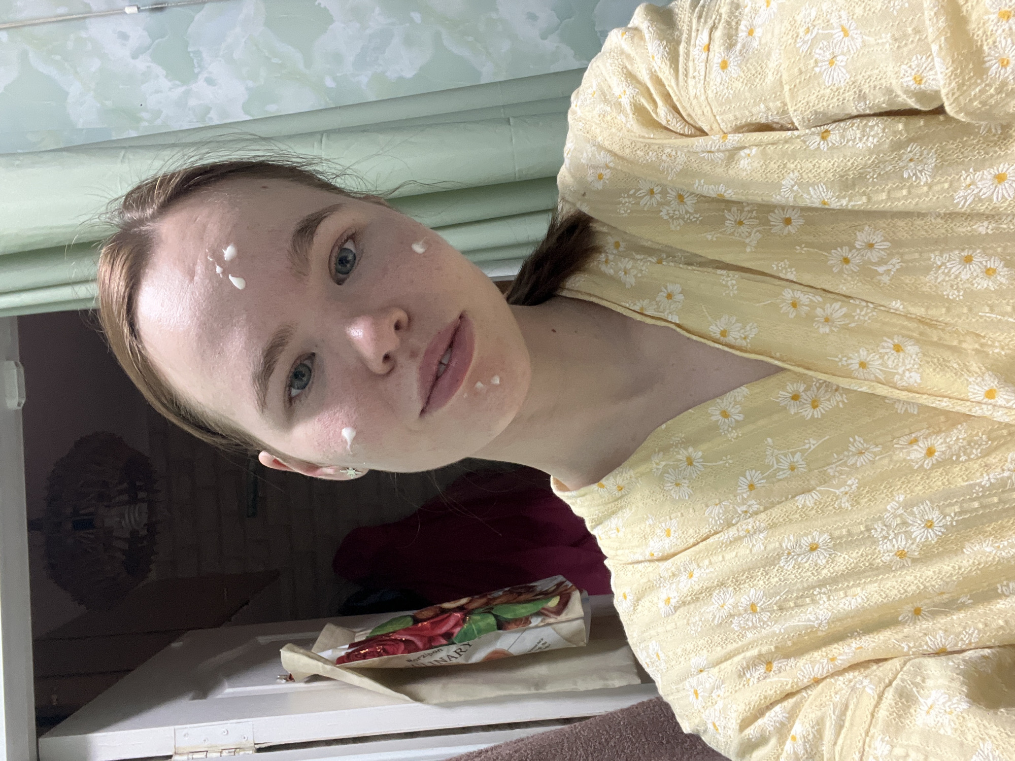 A woman with light skin and brown hair is positioned indoors. She has dabs of white cream on her forehead, cheek, and near her lips. She wears a yellow top with a daisy pattern. Behind her and to the left is a cupboard or cabinet with a package labeled "Marzipan" visible. The setting appears to be Chaikovsky, Russia.