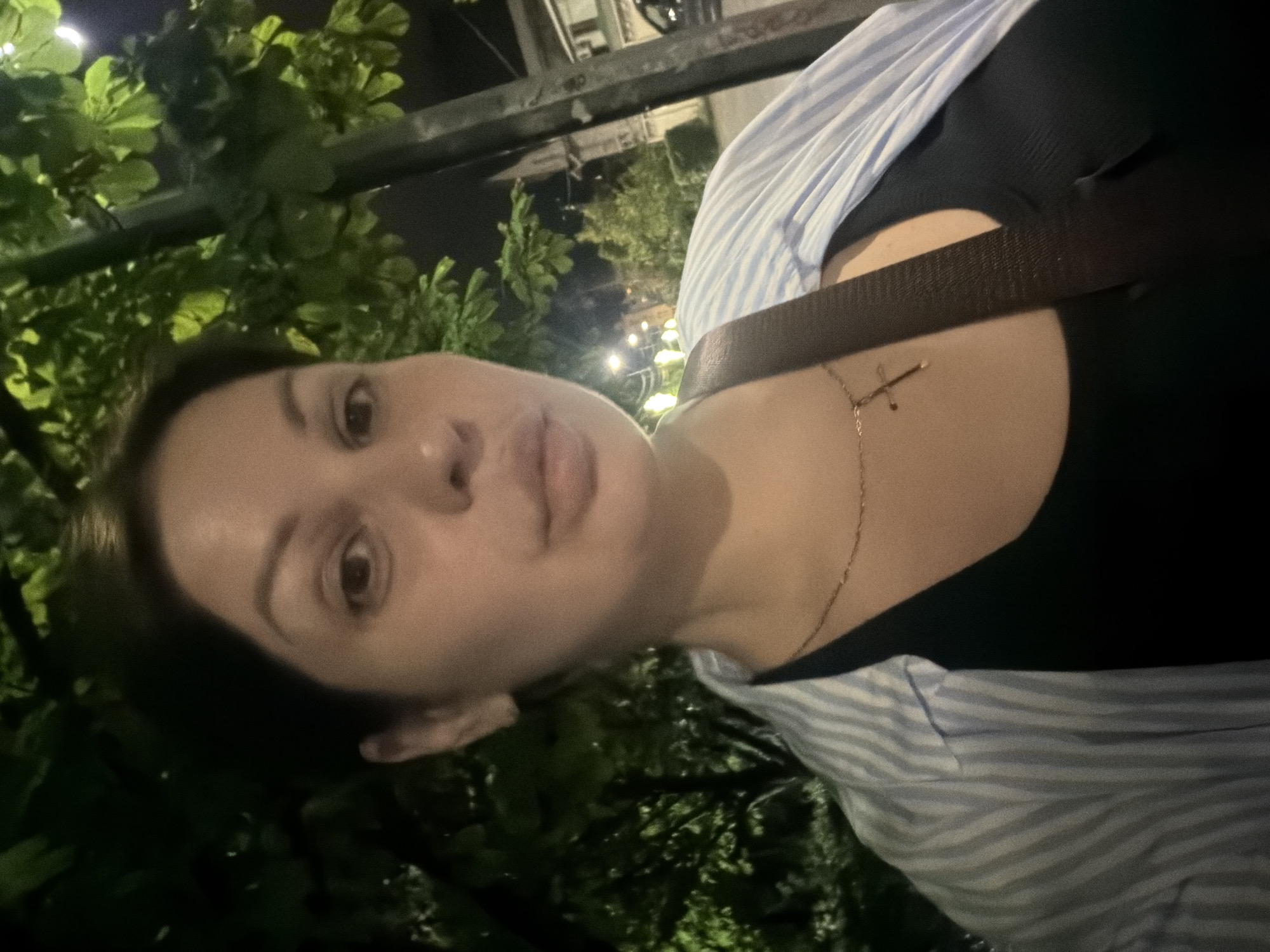 A selfie is taken outdoors at night, featuring a woman with dark hair wearing a light blue and white striped unbuttoned shirt over a black tank top. A thin gold necklace with a cross pendant is visible. She has a brown strap from a bag crossing her chest. The background consists of dark green foliage from trees, some illuminated by artificial light, suggesting an urban setting. Streetlights and buildings are faintly visible in the distance, hinting at a city environment. The image does not contain any information indicating the location is Sochi, Russia.