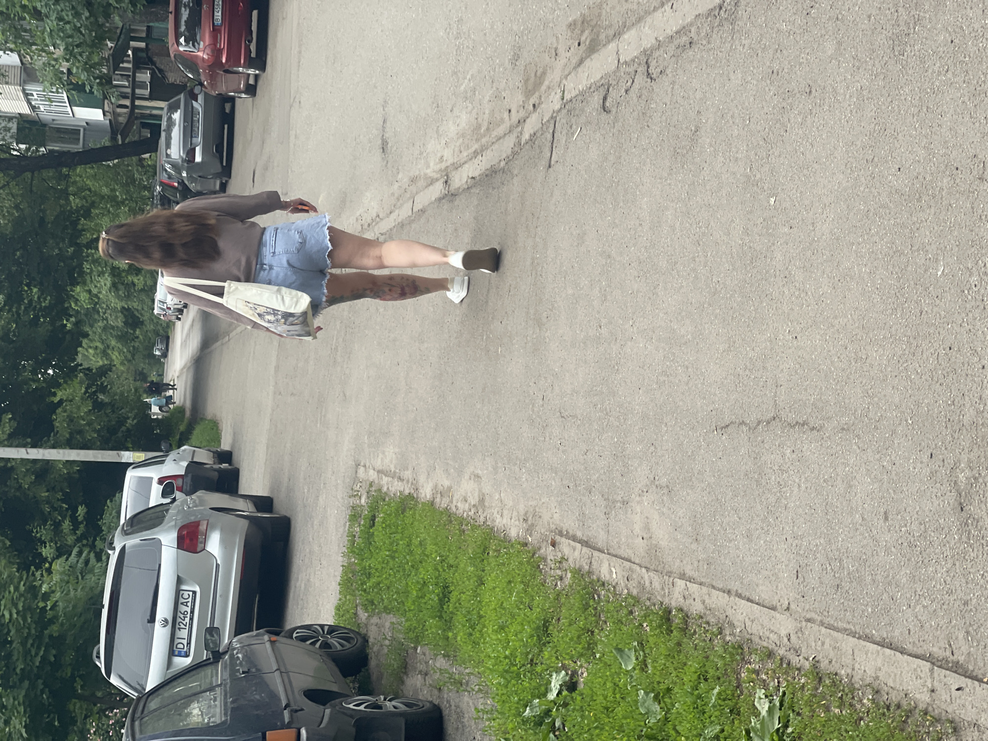 An outdoor, slightly angled shot captures a woman walking away from the camera on a paved path or road. She has long, dark brown hair and is wearing a light brown long-sleeved top, light blue denim shorts, and white sneakers. A light-colored tote bag with a graphic design is slung over her shoulder. Tattoos are visible on her left leg, extending from her calf upwards.
To the left of the path, a strip of green grass and foliage runs alongside parked cars. Several vehicles are visible, including a silver Volkswagen wagon with the Ukrainian license plate "DI 1246 AC" and a dark-colored car in the foreground with distinct multi-spoke wheels. Further down the street, more cars are parked, and parts of buildings are visible through dense green trees in the background. Another red car is partially visible at the top right, with a license plate that starts with "B 4584".
The scene appears to be an urban or suburban street, likely residential, during daytime. The lighting suggests an overcast but bright day, with soft shadows. The license plate "DI" confirms the location as Poltava Oblast in Ukraine, consistent with the provided context of Poltava. No strong emotions are discernible from the woman's back.