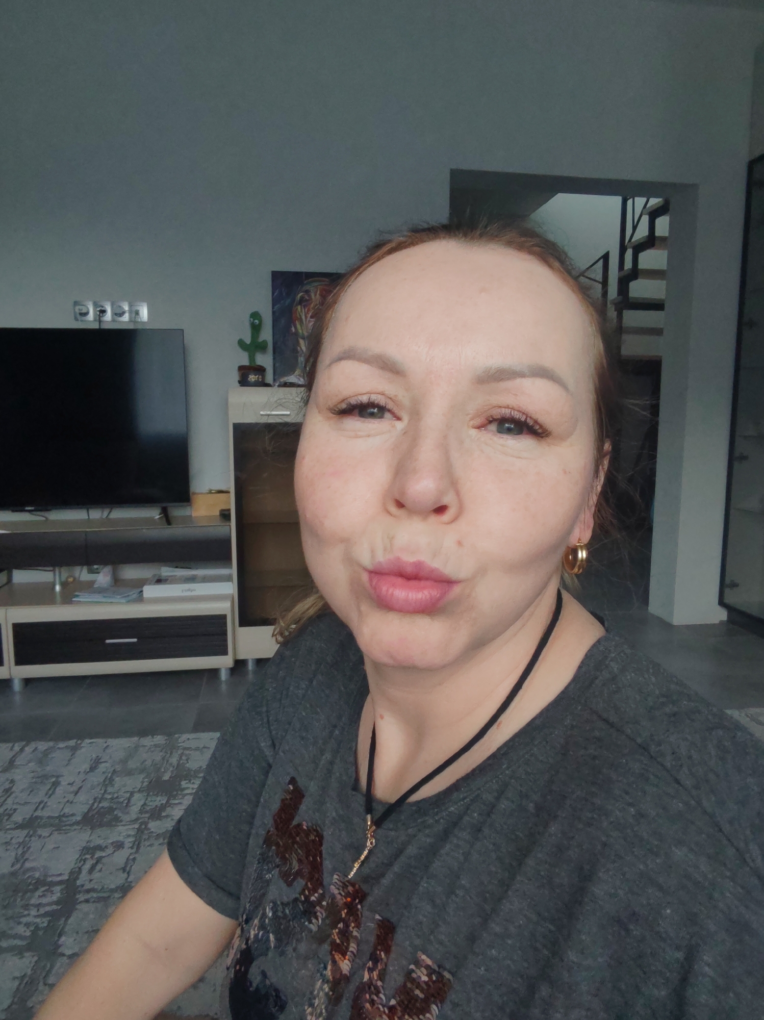 A woman with reddish-blonde hair and fair skin is posing for a selfie indoors. She is wearing a grey T-shirt with brown and black decorative patches and a thin black necklace with a golden pendant. She has gold hoop earrings and is making a pouting expression.
Behind her is a living room setting. A large black television stands on a light-colored entertainment center with brown drawers. To the right of the entertainment center is a tall cabinet with glass doors. A staircase is visible through a doorway in the background. A modern art piece sits on the cabinet. The flooring appears to be gray, possibly tile or carpet with a textured pattern.
The location is Chaikovsky, Russia.