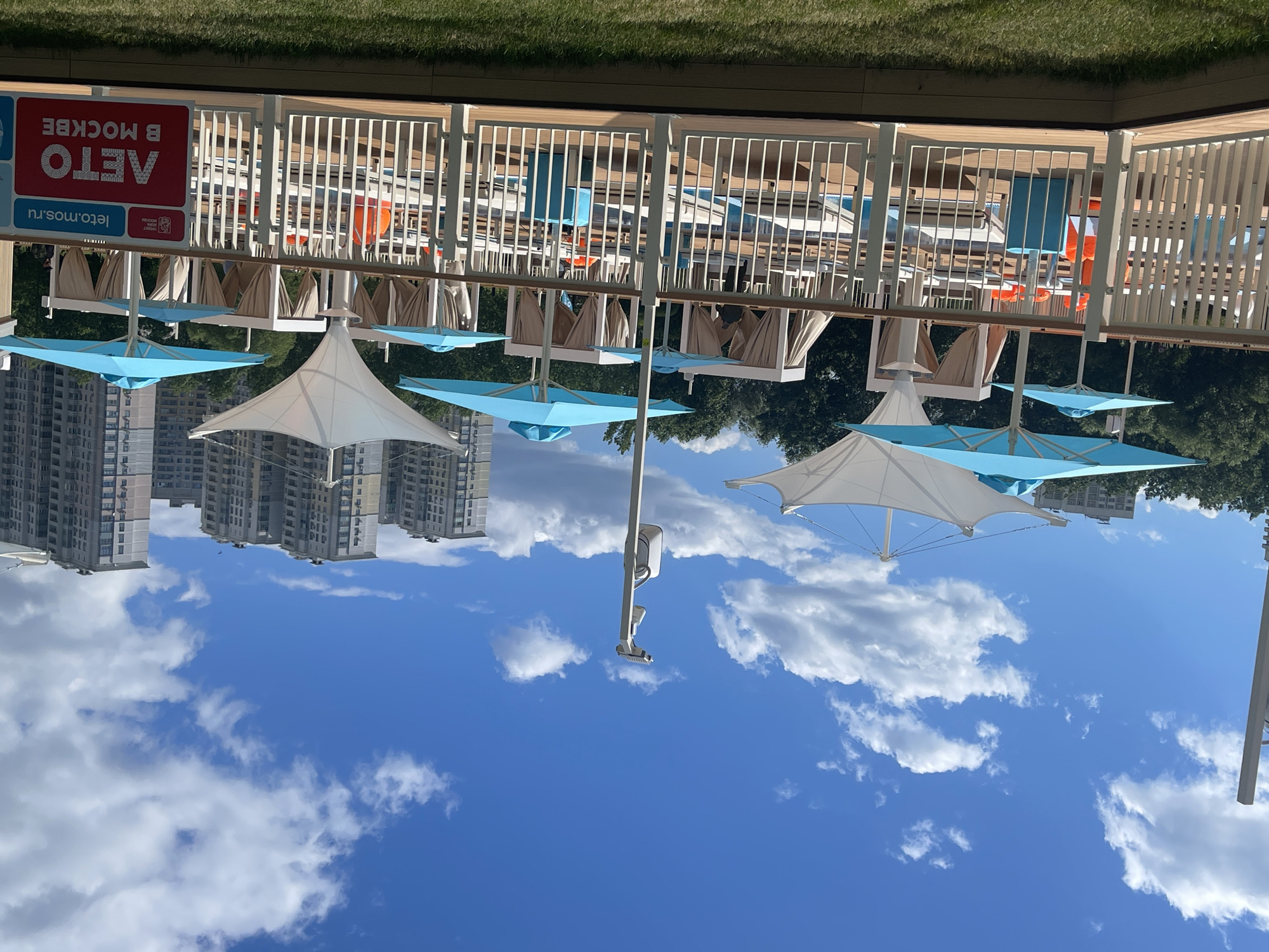 This image captures a sunny day at a resort or outdoor recreational area, likely in Moscow, Russia, as indicated by the Cyrillic text. The scene is dominated by a bright blue sky with scattered white clouds, suggesting fair weather. In the foreground, several large, white and light blue canopy umbrellas are deployed, providing shade. These umbrellas are supported by poles and appear to be part of a seating area with lounge chairs.
Behind the umbrellas, there is a white wooden fence bordering a deck or elevated platform. Beyond the fence, rows of lounge chairs are visible, some with beige canopies. There are also glimpses of orange elements, possibly safety buoys or decorative items, within this area. In the background, a dense line of green trees is present. Further in the distance, tall, multi-story residential buildings are visible, characteristic of an urban environment. The perspective of the image is unusual, with the sky appearing at the bottom and the ground-level elements at the top, suggesting the photo was taken with the camera oriented upside down or the image has been rotated.
A sign is visible on the left side, featuring red and blue sections with white text. The text appears to be in Russian and includes "ЛЕТО" (Summer) and "В МОСКВЕ" (in Moscow), along with a website address "leto.mos.ru". This confirms the location and the likely theme of the area as a summer destination within Moscow. There are no people clearly visible in the image, but the setup implies a place for relaxation and leisure. The lighting suggests it is daytime, likely late morning or early afternoon.