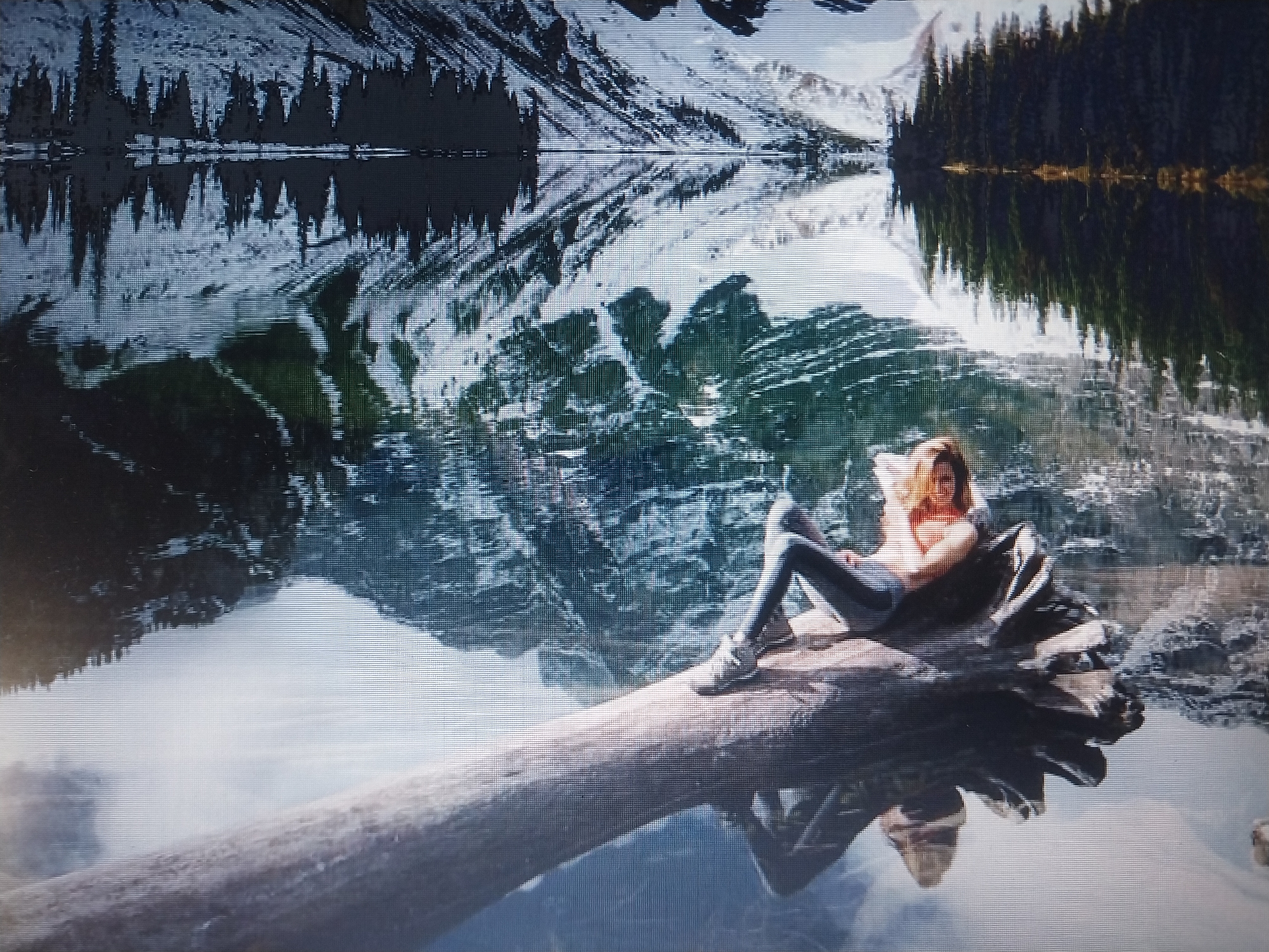 A woman is lying on a log that is partially submerged in a calm body of water. The log is in the foreground, stretching diagonally across the image from the bottom left to the middle right. The woman is lying on her side, propped up on her elbow, with her legs bent. She is wearing a light-colored top and dark pants. Her hair is light brown and shoulder-length. Behind the log and the woman, the water reflects a picturesque landscape. This landscape consists of snow-capped mountains, dark green coniferous trees lining the shore, and a clear sky. The reflection is very clear, creating a symmetrical image with the real landscape. The overall mood is serene and tranquil.