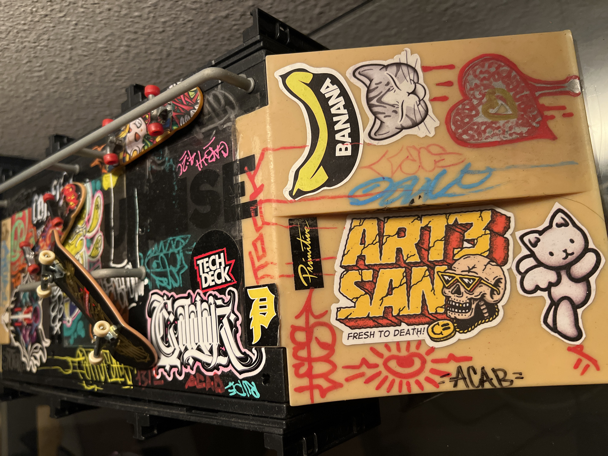 The image shows a close-up, high-angle view of a fingerboard skate park setup. The park is adorned with numerous stickers, including graffiti-style designs, brand logos such as "Primitive" and "Tech Deck," and various cartoonish images like a banana, a cat, a skull, and a winged teddy bear. Two fingerboards are visible on the park, one in the foreground with a black and gold design, and another further back with a colorful graphic. The skate park appears to be constructed from plastic and has metal rails. The background is out of focus and shows a textured wall. The scene is well-lit, with the focus sharply on the fingerboard park and its detailed stickers. There is no information in the image that suggests a location in Madrid, Spain.