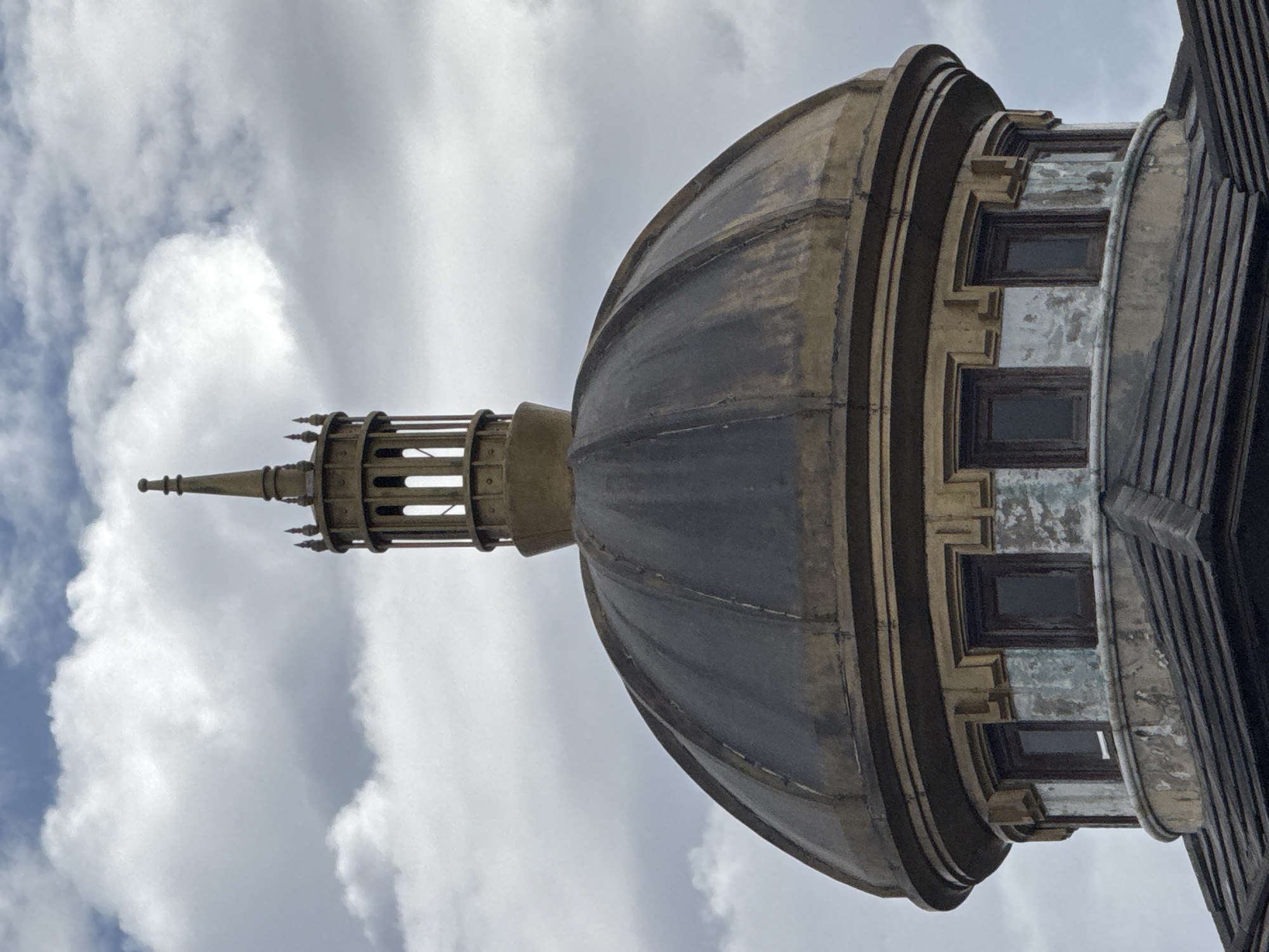 The image shows the dome of a building against a cloudy sky. The dome is made of dark metal with golden trim and has a lantern at its apex. Below the dome, there is a circular section with several small, dark windows set into it. This section appears weathered, with peeling paint in patches of blue and white. A dark, tiled roof structure is visible beneath the circular section. The building's architecture suggests it might be a historical or government building. The image does not provide specific details about the city of Da Lat, Vietnam.