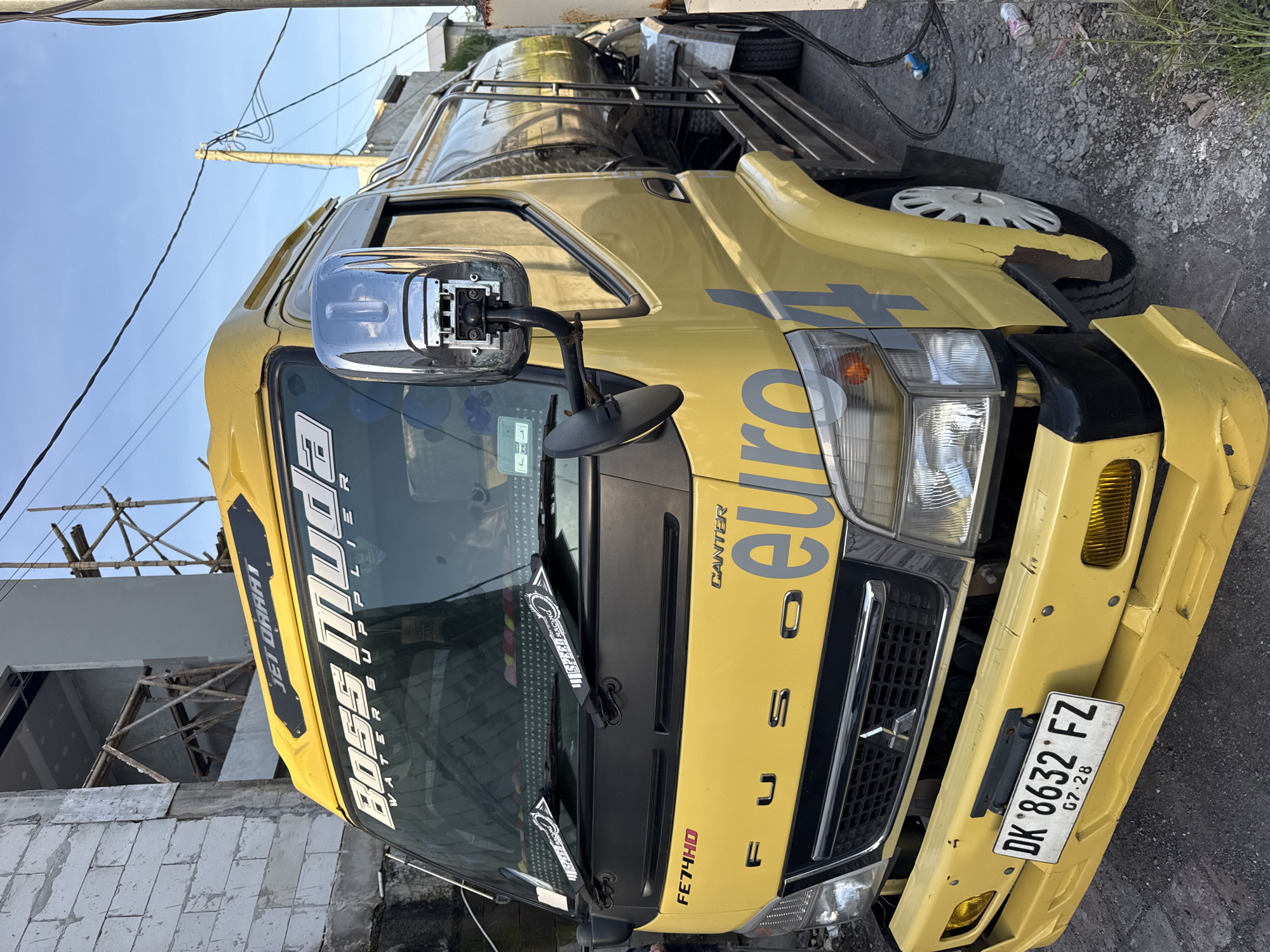A yellow Fuso Canter truck, specifically an FE74HD model with "euro4" decal, is parked on an asphalt road. The truck's license plate reads "DK 8632 FZ". A partially constructed building with bamboo scaffolding is visible in the background. The location is Kuta Selatan, Indonesia. The image shows a vehicle on a street in a residential or commercial area. No people are visible.