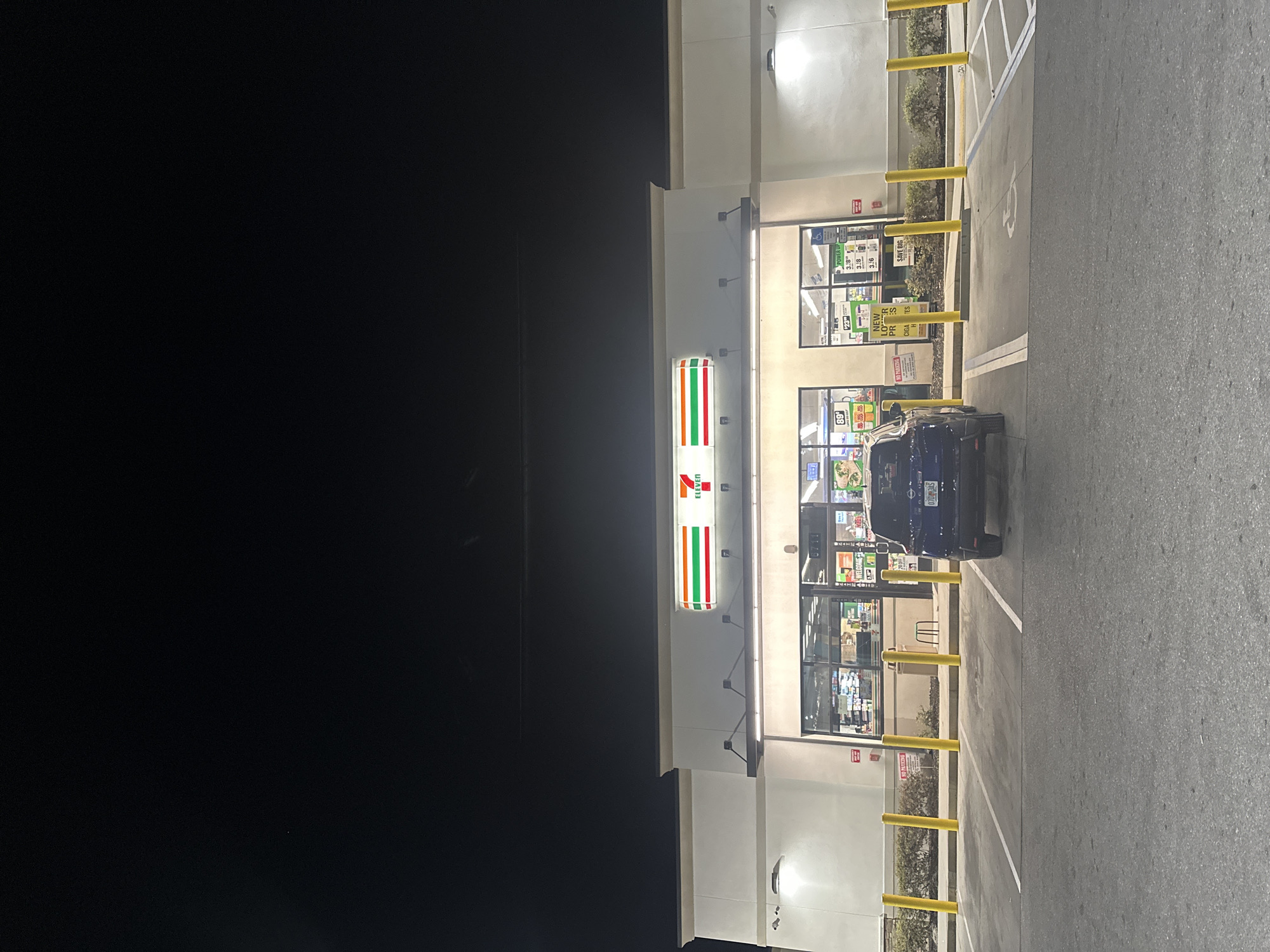 The image shows a 7-Eleven convenience store at night. A dark blue vehicle, likely a car, is parked in front of the store in a parking space. The 7-Eleven signage is prominently displayed above the entrance. The store's front is well-lit, showing product displays and windows. Parking spaces are lined with yellow bollards. The environment appears to be a typical commercial area at night in a location in the city of Homosassa Springs, United States of America.