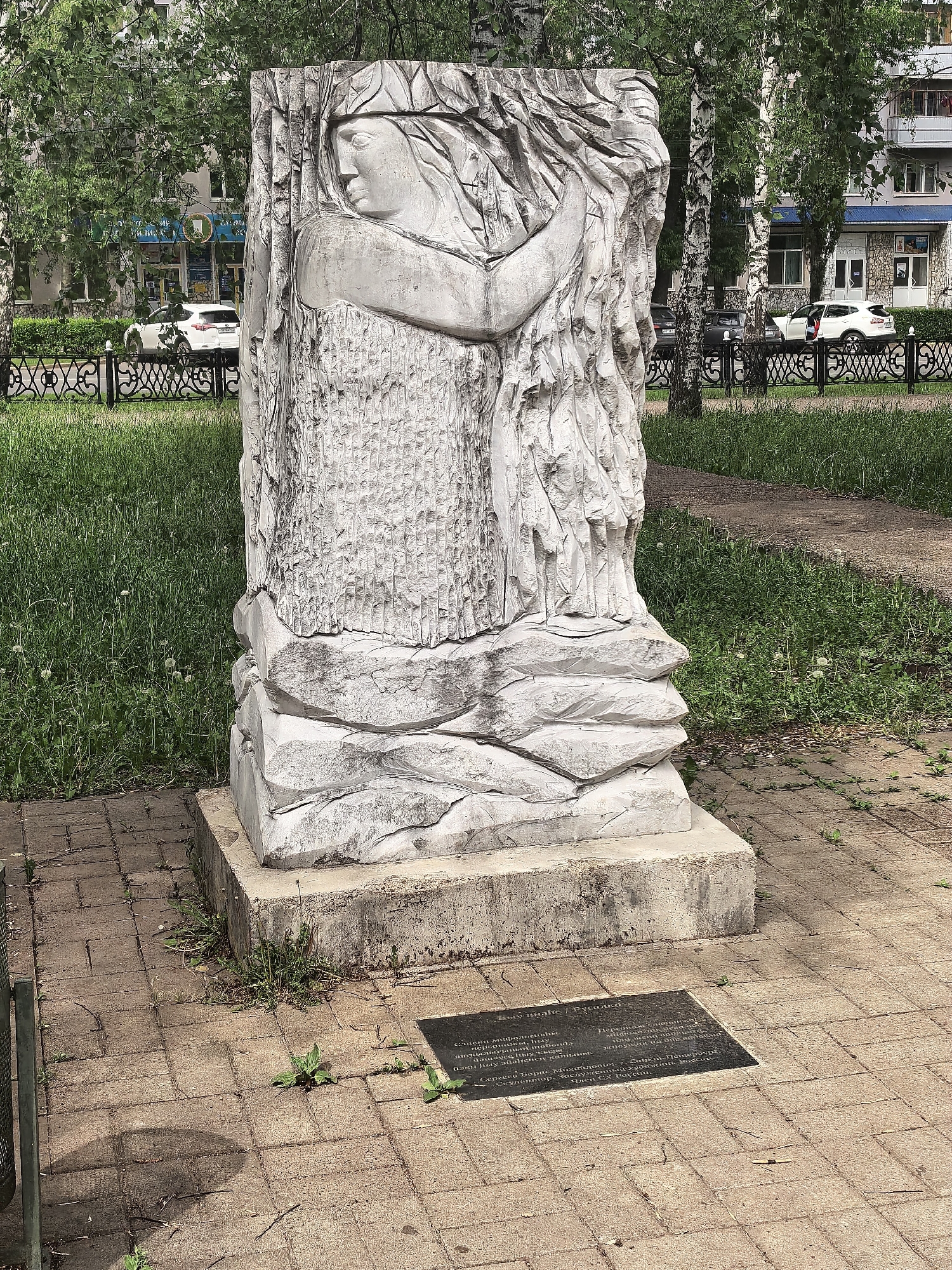 The image shows a sculpture titled "Rusalka" located in Sterlitamak, Russia. The sculpture is made of white stone and depicts a woman with long hair, possibly a mythical water spirit. It stands on a concrete base with a plaque in front of it, containing Russian text. The surrounding environment includes a park-like setting with grass, trees, and a sidewalk paved with brick. A black metal fence and buildings are visible in the background. The photo was taken during daylight hours.