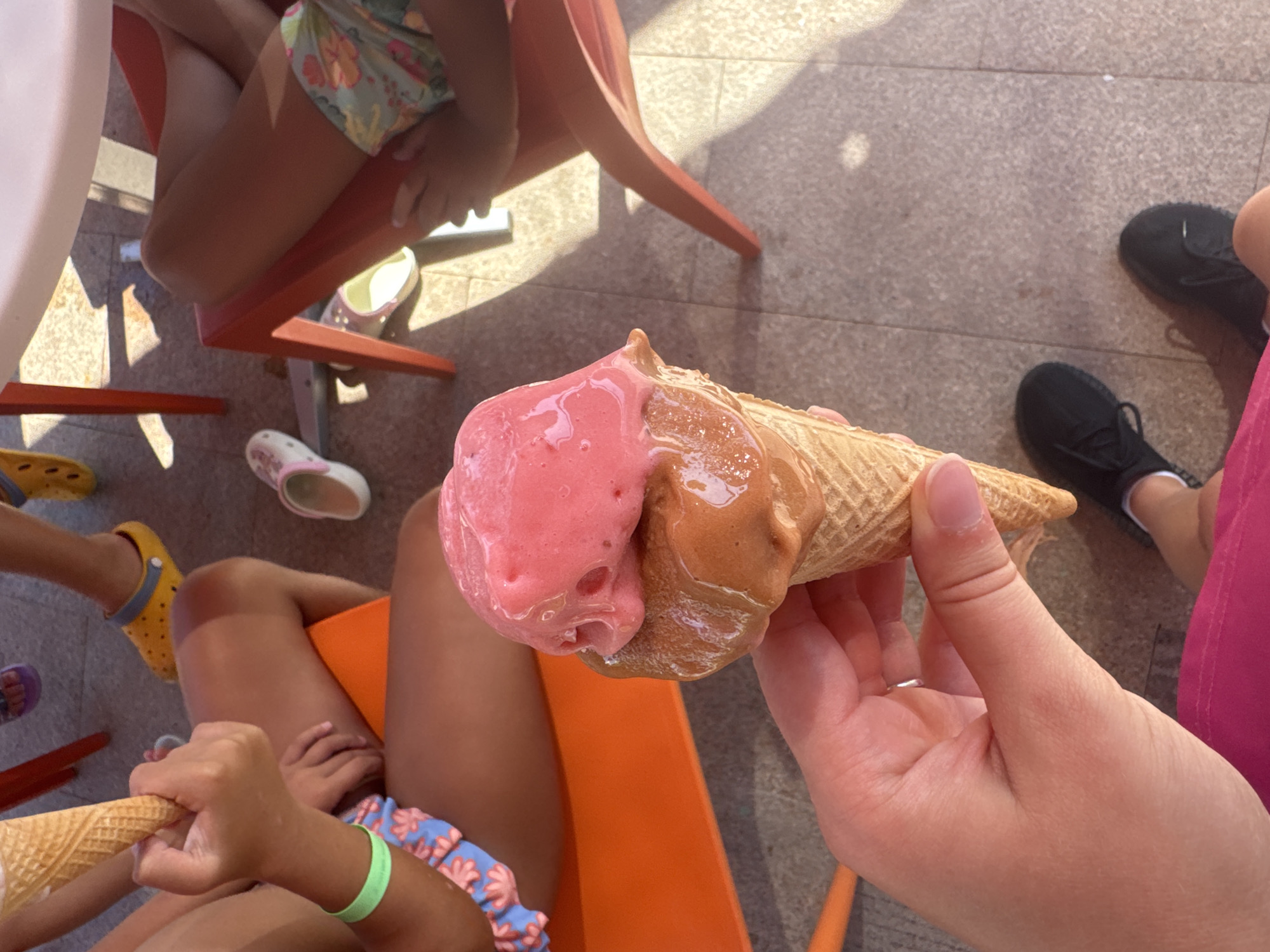 A close-up shot shows a hand holding a double-scoop ice cream cone. The cone has a waffle pattern and is filled with two scoops of ice cream: one strawberry-flavored and the other chocolate-flavored. The ice cream is beginning to melt, with drips visible on the cone. In the background, several children are seated on chairs around a table, their legs and feet visible. One child is wearing yellow Crocs, another a pink sandal, and a third is barefoot. The scene appears to be outdoors on a tiled surface, with sunlight casting shadows. The location is identified as Aksu, Turkey, suggesting a warm climate, possibly a park or outdoor eating area.