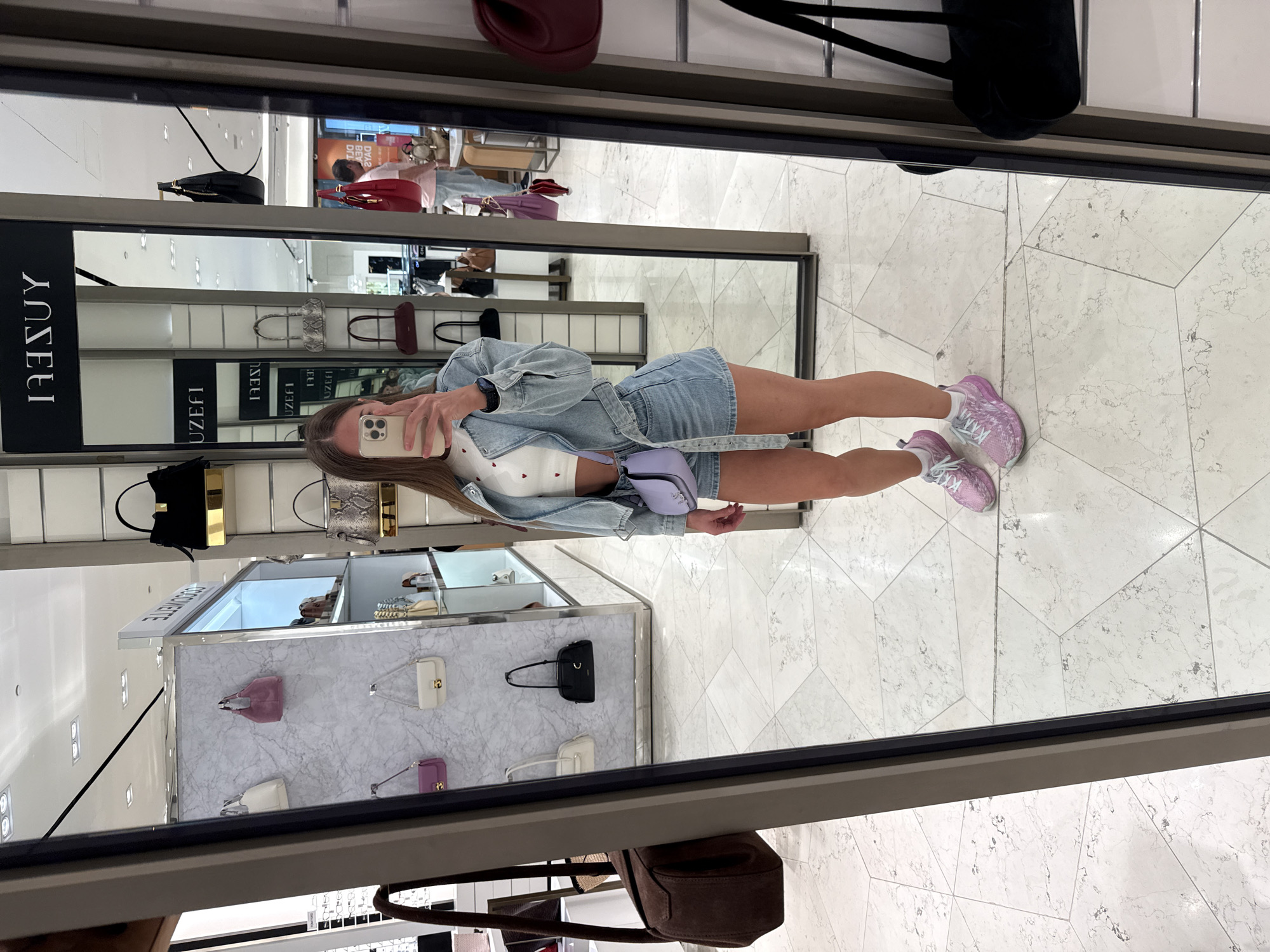 A young woman is taking a mirror selfie in what appears to be a high-end department store in Saint Petersburg, Russia. She is wearing a denim jacket, a white cropped top with red heart patterns, and denim shorts. She is also carrying a light purple crossbody bag and wearing pink athletic shoes. The background shows displays of handbags on shelves and mannequins, as well as a man in the background browsing merchandise. The floor is tiled with a geometric pattern. The store appears to be well-lit and modern.