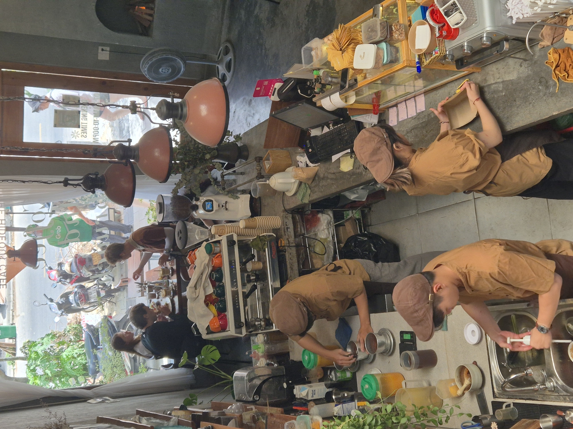 The image depicts the interior of a coffee shop, likely in Đà Nẵng, Vietnam, as suggested by the presence of a Grab delivery person, a common sight in Vietnamese cities. The viewpoint is from above, looking down into the cafe.
In the foreground, a young person wearing a brown cap and brown shirt is washing dishes in a stainless-steel sink. They are actively pumping soap into the sink. To the right of the sink, a person in a similar brown outfit and cap is preparing packaging materials, holding a cardboard container and a yellow cutting tool. Behind them, an espresso machine is visible, along with stacks of paper cups and various bottles and containers.
Further back, a person wearing a brown cap and shirt is seen from the side, working at a counter and appearing to prepare a beverage. They are holding a small metal cup and a silver device.
In the midground, a group of three people are seated at a table. Two individuals are eating, and another person is looking towards them. This area is near a large window that looks out onto a street with parked motorcycles and a person in a green Grab delivery uniform.
The cafe interior features industrial-style lighting fixtures with copper-colored shades, potted plants, and exposed concrete elements. The overall atmosphere appears to be busy and functional, with staff actively engaged in their tasks and customers enjoying their time. The scene is lit by a combination of natural light from the window and artificial lighting from the overhead lamps.