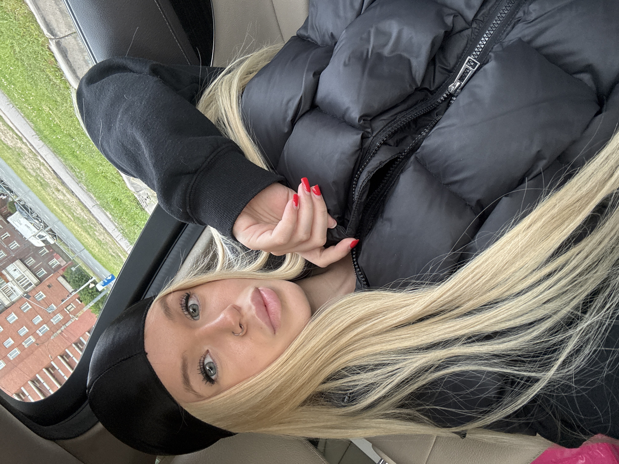 The media file is a close-up, rotated portrait of a young woman with long blonde hair and blue eyes, wearing a black puffer jacket and a black beanie. She is looking at the camera with a neutral expression. A silver nose ring is visible. Her nails are painted red. Her left hand is resting on her jacket, with her fingers slightly curled. The setting appears to be inside a car, with the window showing a view of a brick building and some greenery. The lighting suggests it is daytime. There is no visible text in the image.