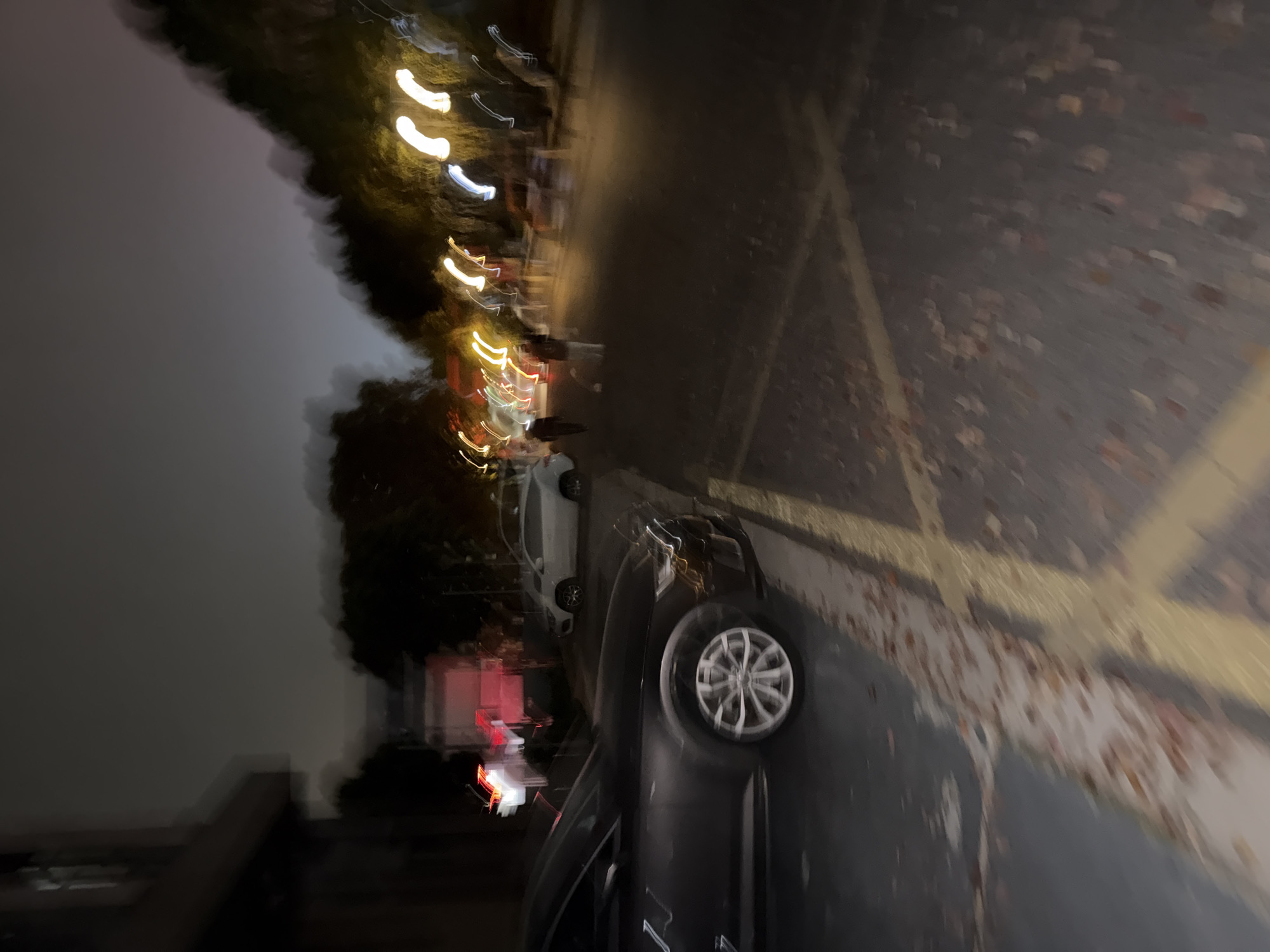 The image presents a blurry nighttime scene, likely captured with camera movement. In the foreground, a black car is parked along a road with marked parking spaces in yellow paint. The pavement is littered with what appears to be fallen leaves. Across the road, blurred lights, possibly from streetlights and other vehicles, indicate a developed area. Hints of trees are visible, suggesting a street lined with vegetation. Far in the background, there is a white building with a red sign. While the image suggests an urban environment, the specific details confirming the location as Jiangning District, China, are not visually evident.