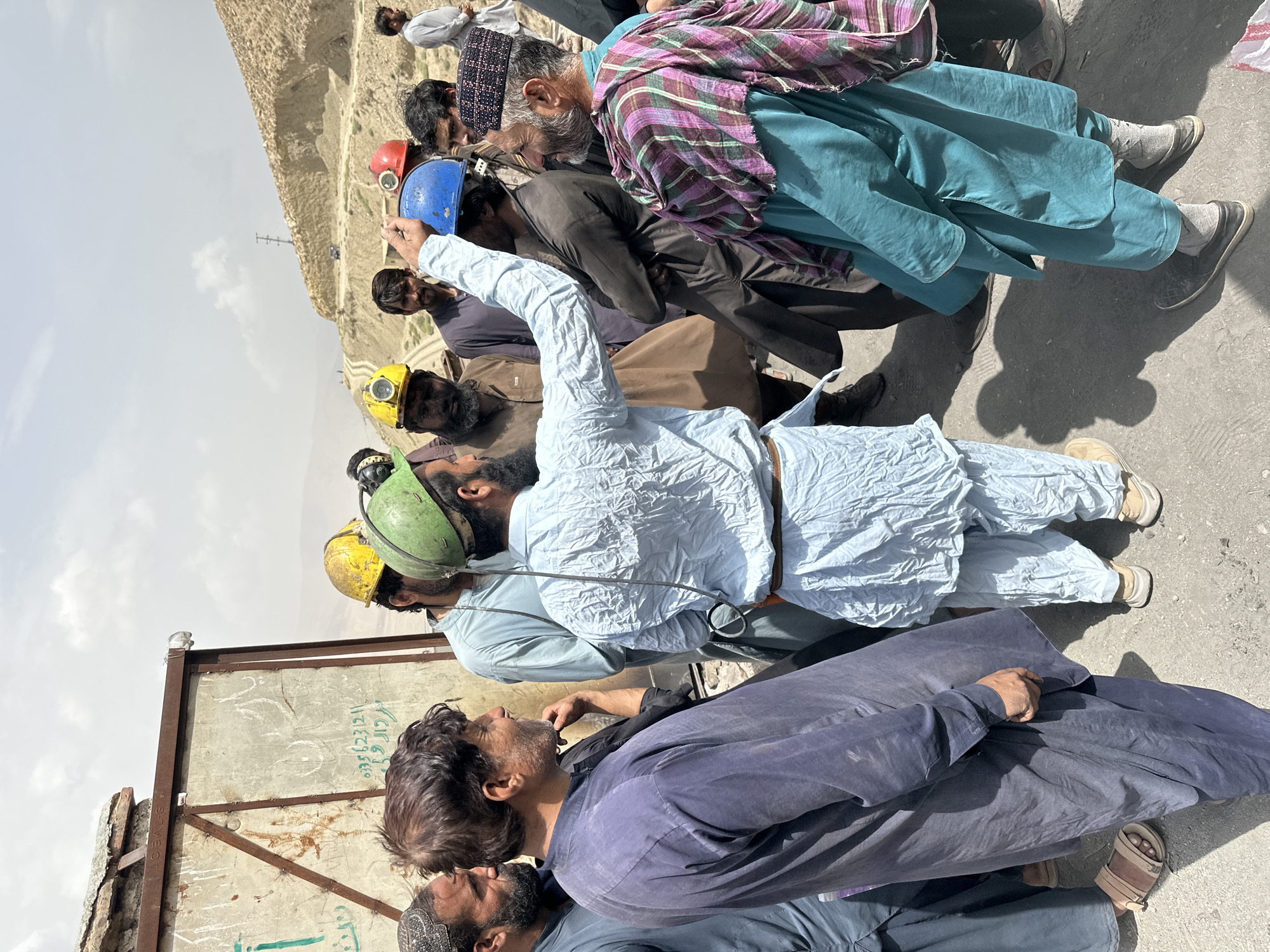 A group of men, some wearing hard hats, are gathered outdoors in what appears to be a dusty, mountainous region, possibly a mining area. The men are dressed in traditional clothing, including shalwar kameez. One man in a light blue outfit is gesturing and speaking to the others, who are listening attentively. The environment suggests a harsh, arid climate with dry, rocky terrain and a visible dirt road leading up a hillside. The presence of construction materials or debris and writing on a corrugated metal structure in the background might indicate an industrial or work site. The overall atmosphere is one of serious discussion or briefing. The location is identified as Quetta, Pakistan.