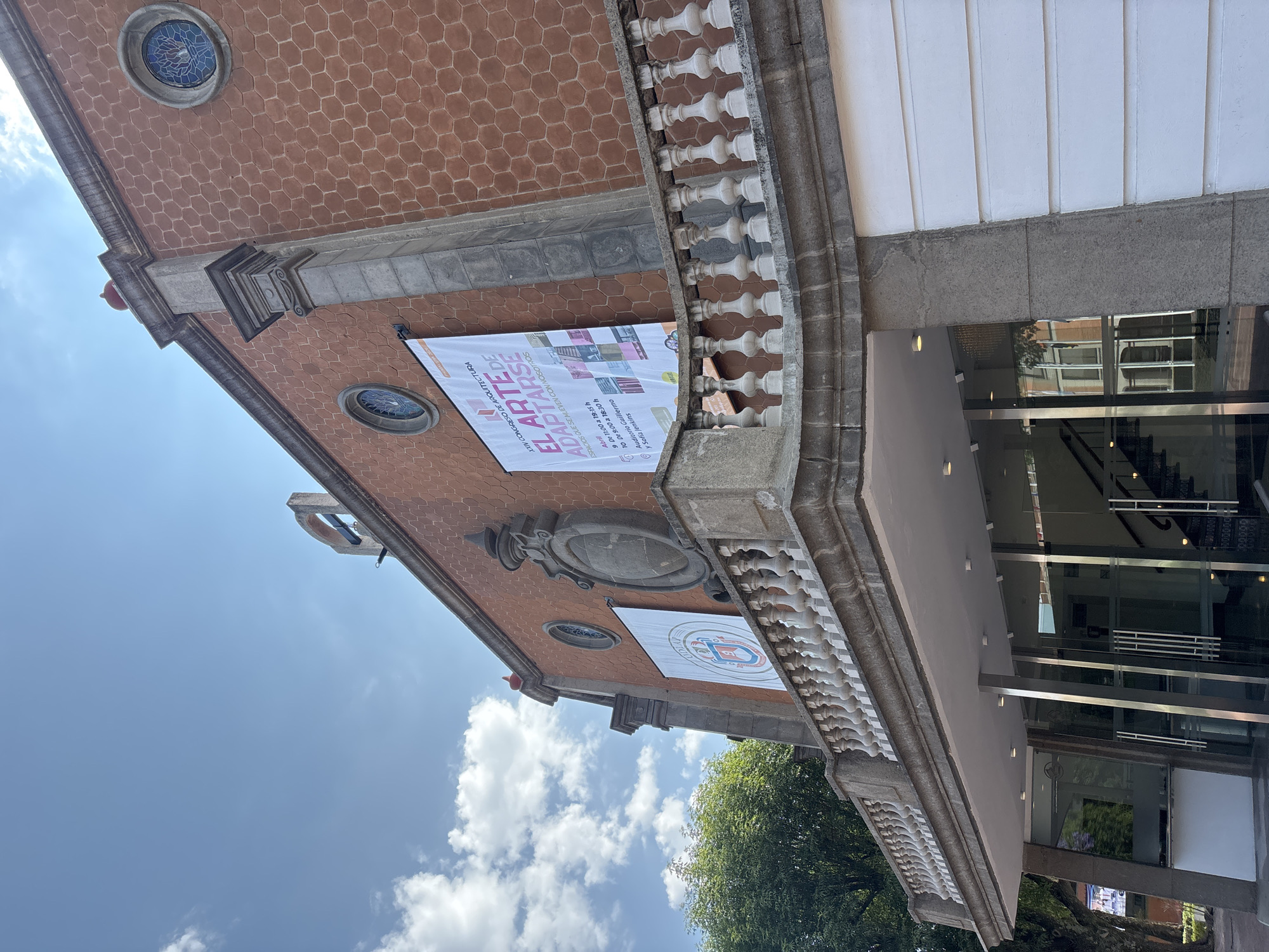 The image depicts the exterior of a building in San Andrés Cholula, Mexico. The building has a red-brown hexagonal patterned facade, accented with decorative stone elements. Two banners hang from the building, one advertising the XXIV Congress of Architecture, featuring the phrase "El Arte de Adaptarse." The other banner displays a crest. A white balustrade runs along the edge of the building. The lower part of the building includes a glass-fronted entrance with a canopy. The sky is visible with scattered clouds. Part of a tree is visible in the bottom-right corner.