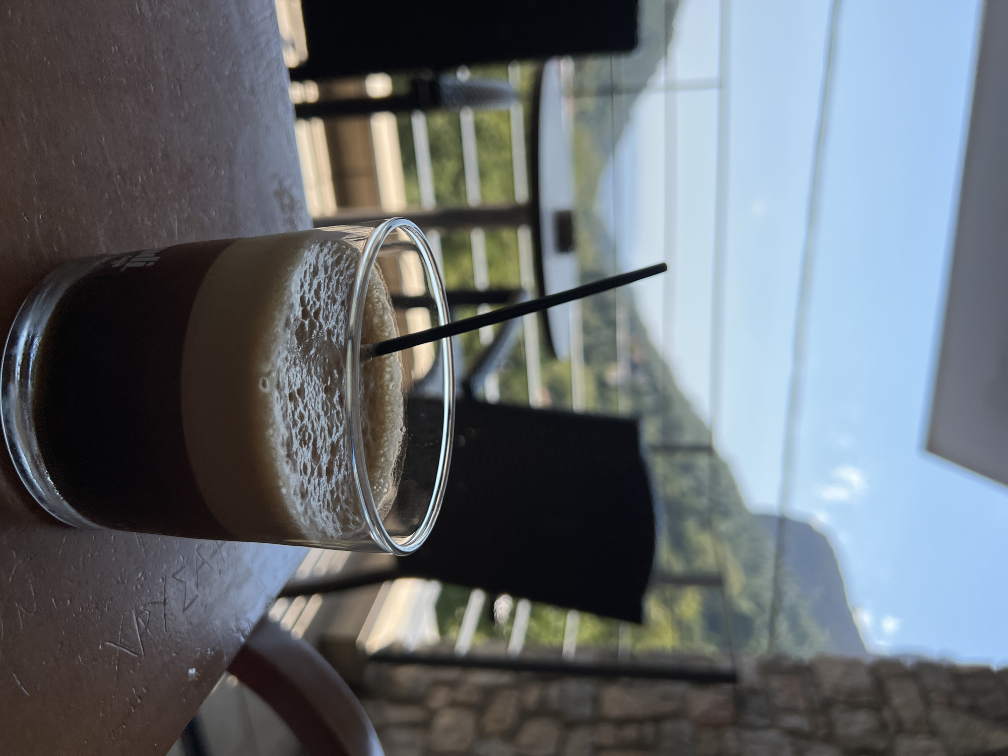 A close-up shot focuses on a glass of iced coffee with a black straw on a wooden table. The coffee is layered, with a dark bottom and a foamy top. In the background, through a railing, a scenic view of a valley with lush green trees and mountains is visible under a clear blue sky. The location appears to be an outdoor cafe or restaurant, possibly in the village of Stení Dirfyos, Greece, given the mountainous landscape. The image is taken from a slightly low angle, with the coffee glass being the primary subject.
