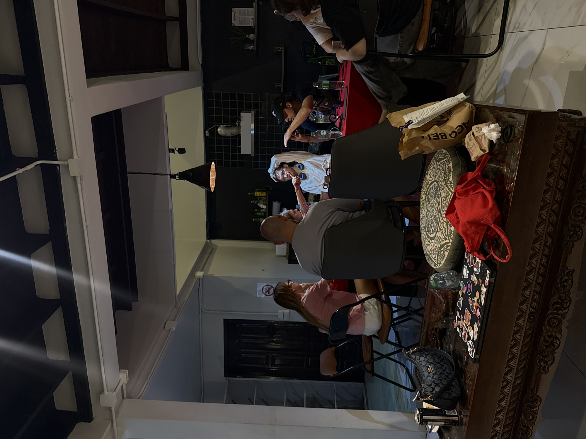 The image depicts an indoor gathering of six individuals seated around a red table. A woman in a pink shirt and white shorts is on the left, looking away. Next to her, a bald man in a grey t-shirt is also looking away. In the center, a person wearing glasses and headphones is gesturing with their hands, while a man in a black baseball cap is holding a phone and also gesturing. On the right, another person in a white t-shirt is partially visible, and further right, a person in a black t-shirt is seated.
The environment appears to be a room with dark walls and a patterned ceiling. Hanging from the ceiling is a black pendant light. Shelving units are visible in the background, with various items on them, including bottles and what appear to be decorative objects. A black door is also visible.
In the foreground, a dark wooden coffee table is laden with objects, including a black handbag with a gold chain, a laptop covered in stickers, a red fabric item, a paper bag with "TACO BELL" printed on it, and crumpled white paper. A patterned cushion is placed on the floor.
The lighting is dim, with the pendant light and possibly other ambient lights illuminating the scene. The overall atmosphere suggests a casual social gathering. The location is identified as Chiang Mai City Municipality, Thailand.