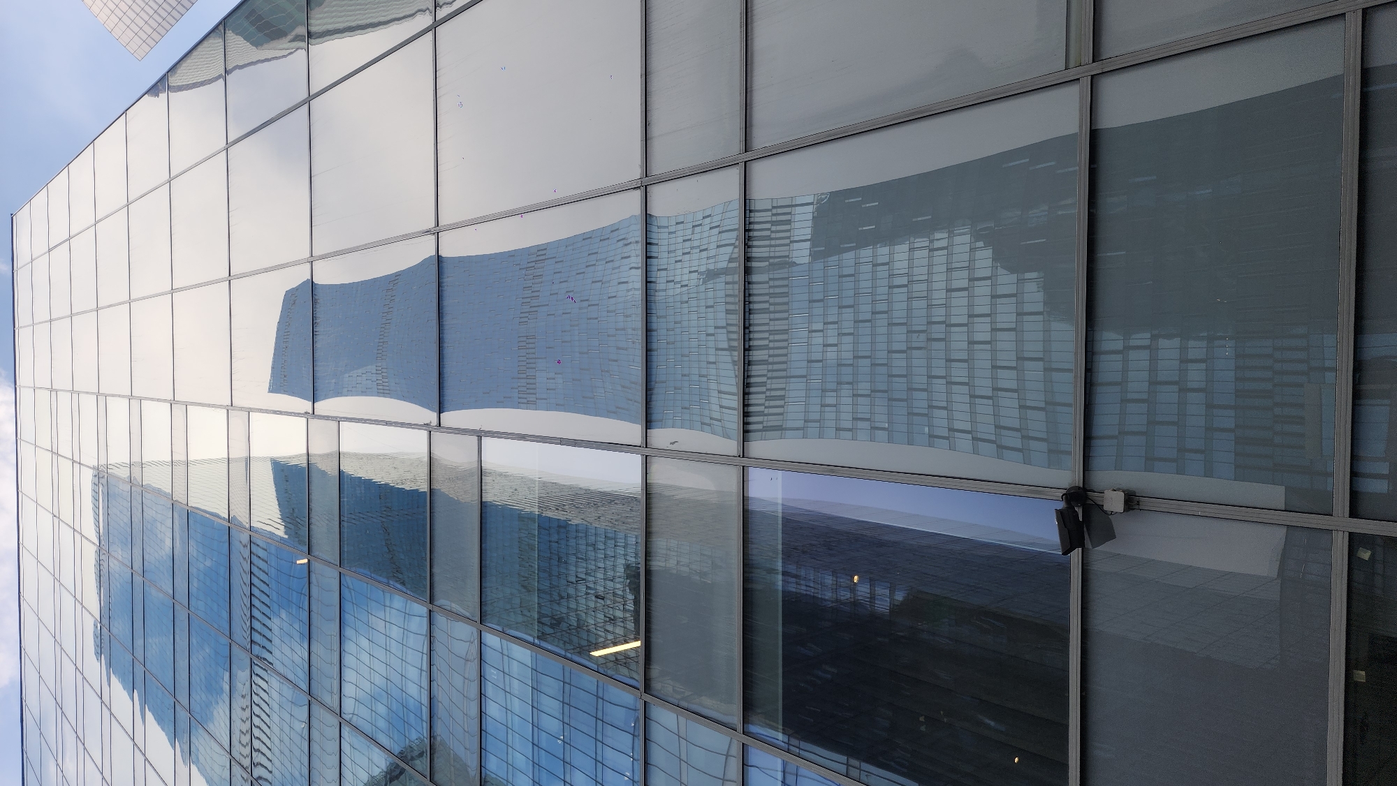 The image depicts the exterior of a modern building with a glass facade, likely located in the city of Moscow, Russia. The building's windows reflect the sky and other structures, creating an illusion of depth and complexity. The reflections are distorted and curved, suggesting that the glass panels are not perfectly flat. The building's structure is evident through the grid-like pattern of the window frames, which are dark in color. There is an electronic device on the side.