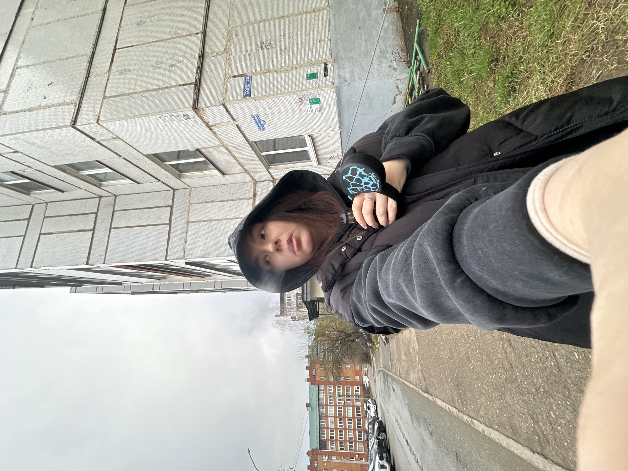 A young woman is taking a selfie outside of a building. The woman has long brown hair and is wearing a black hooded jacket and a grey sweatshirt. She is holding a pair of headphones in her left hand. The building is a multi-story apartment building with white tile on the exterior. There is a street sign on the side of the building. Across the street, there is another multi-story apartment building. Cars are parked along the street. The sky is overcast. This location is Chaikovsky, Russia.