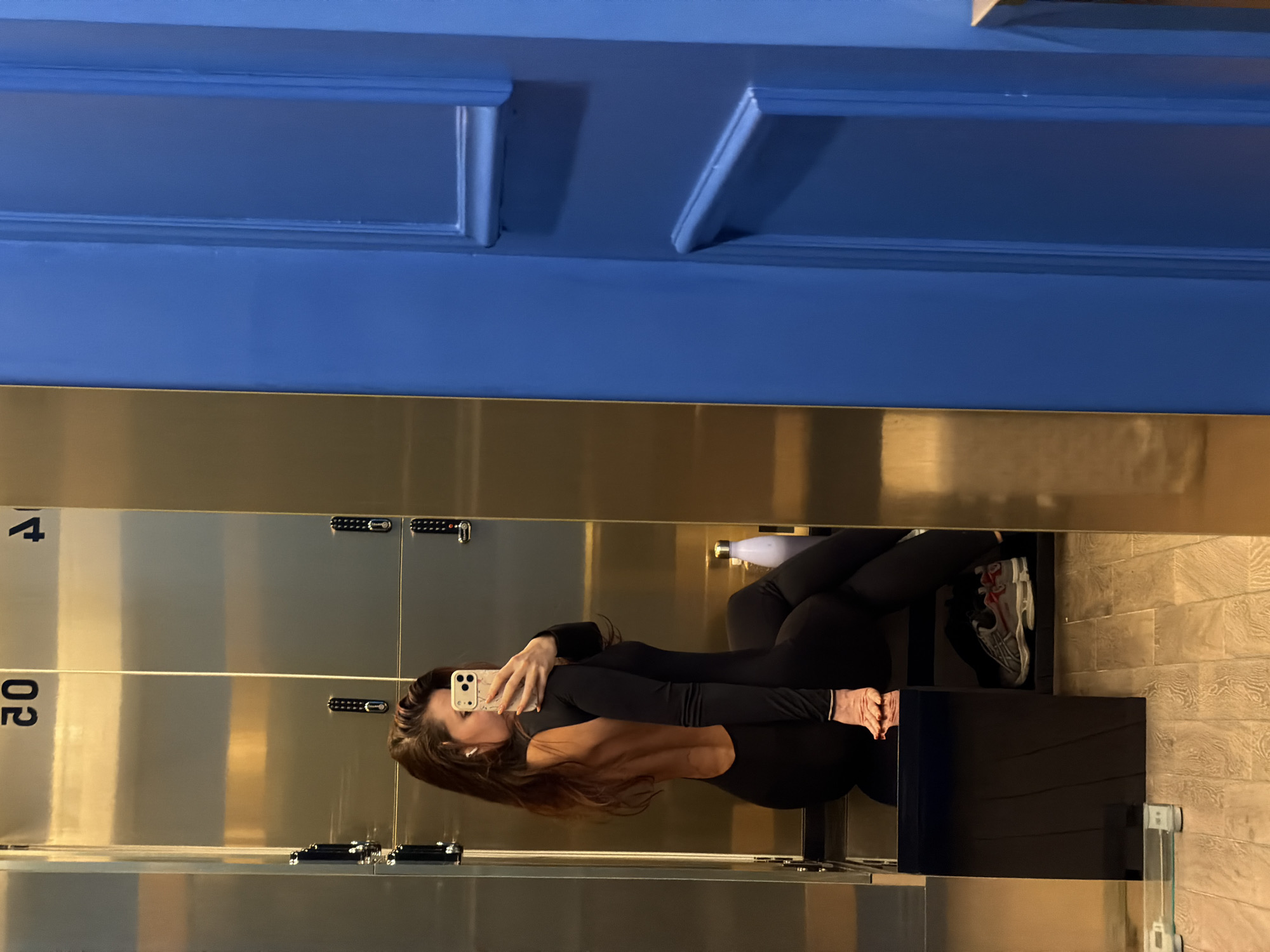 A woman is taking a mirror selfie in a locker room. She is wearing a black jumpsuit and holding an iPhone. The lockers are silver with combination locks. The wall above the lockers is blue. A pair of running shoes and a water bottle are visible behind her.
The image was taken in Belgrade, Serbia.