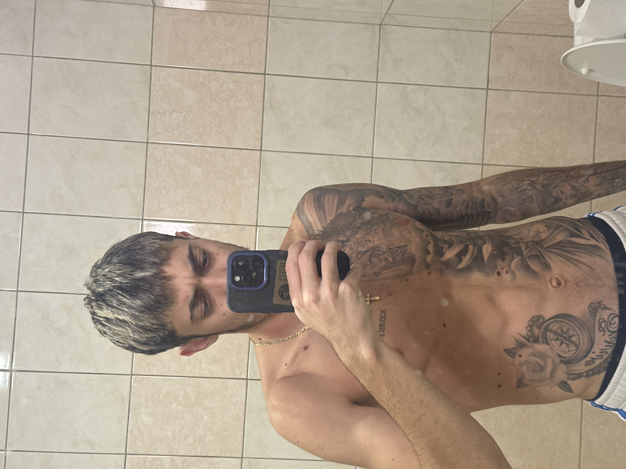 A shirtless man with tattoos and a gold chain takes a selfie in a bathroom. He is holding a dark-colored smartphone with multiple camera lenses. The bathroom has beige square tiles. The image does not depict a location in Varna, Bulgaria.