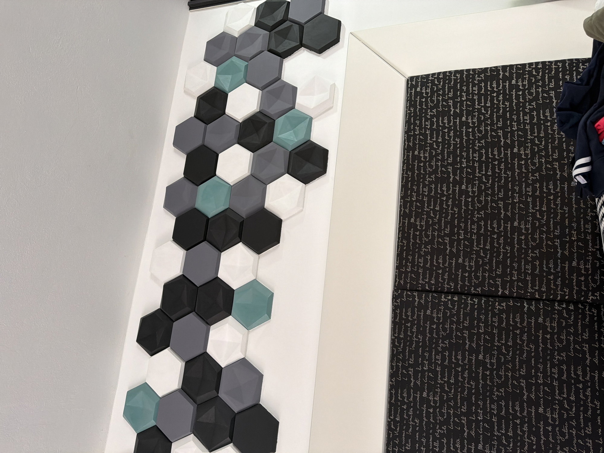 The image shows a wall decorated with hexagonal tiles in black, gray, white, and teal colors, arranged in a vertical pattern. To the right of the tiled wall is a surface covered in a black and white patterned fabric featuring handwritten-style text. Some clothing can be seen draped to the right, obscuring some of the patterned fabric. The environment appears to be indoors, possibly a room in a home or building. The location is unknown and cannot be confirmed as Puebla, Mexico.