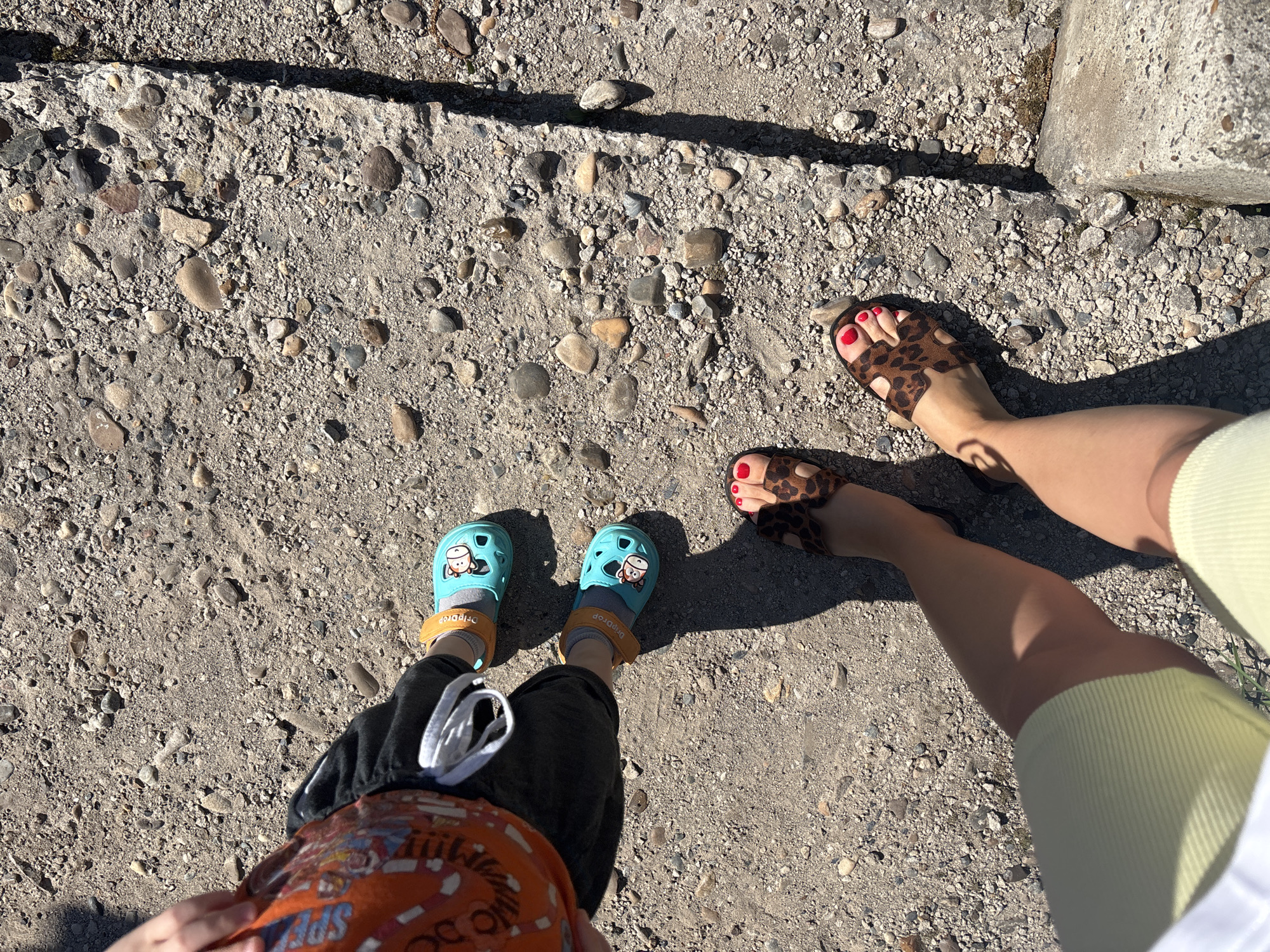 The image shows a top-down view of two individuals standing on a gravel-like surface with some larger stones interspersed.
The person on the left, presumably a child, is wearing black shorts, an orange shirt with graphics, and blue Crocs-style shoes with a cartoon character. The child also has grey socks.
The person on the right is wearing a light-yellow skirt or shorts and leopard-print sandals. They have red nail polish on their toes.
The ground is a mix of small gravel and larger stones. The lighting suggests it is daytime, and shadows are visible. A concrete border is visible on the right side of the frame.
The exact location within the city of Ферма, Russia is not discernible from the image. There is no other contextual information to determine actions or interactions.