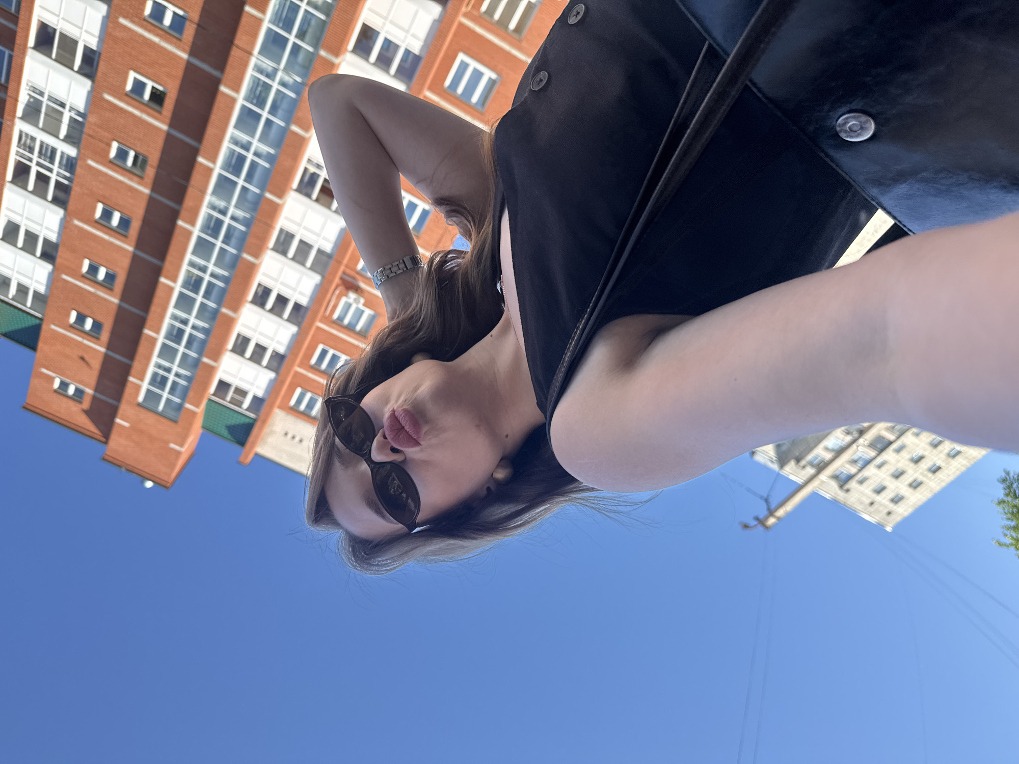 A low-angle, wide shot captures a young Caucasian woman from the chest up, standing outdoors on a clear, sunny day. She is positioned in the foreground, looking upwards towards the camera with her head tilted back. The woman has long, light brown hair, is wearing dark sunglasses, and has her lips pursed in a duckface gesture. She is dressed in a black sleeveless top, adorned with two circular buttons on the front. Her right arm is raised and bent, with her hand resting on the back of her head, and she is wearing a silver-colored watch on her left wrist. A dark-colored bag is slung over her right shoulder.
Behind her, dominating the upper half of the frame, is the facade of a multi-story brick building, likely residential, with numerous windows. The building is oriented at an angle, suggesting the photo was taken from a street level perspective, looking up. The lower section of the image reveals a section of another building with a lighter facade, and a telephone pole with visible wires. The sky is a bright, cloudless blue. The overall scene is indicative of a typical urban environment, likely within the city of Chaikovsky, Russia, given the architectural style of the buildings and the presence of such infrastructure.