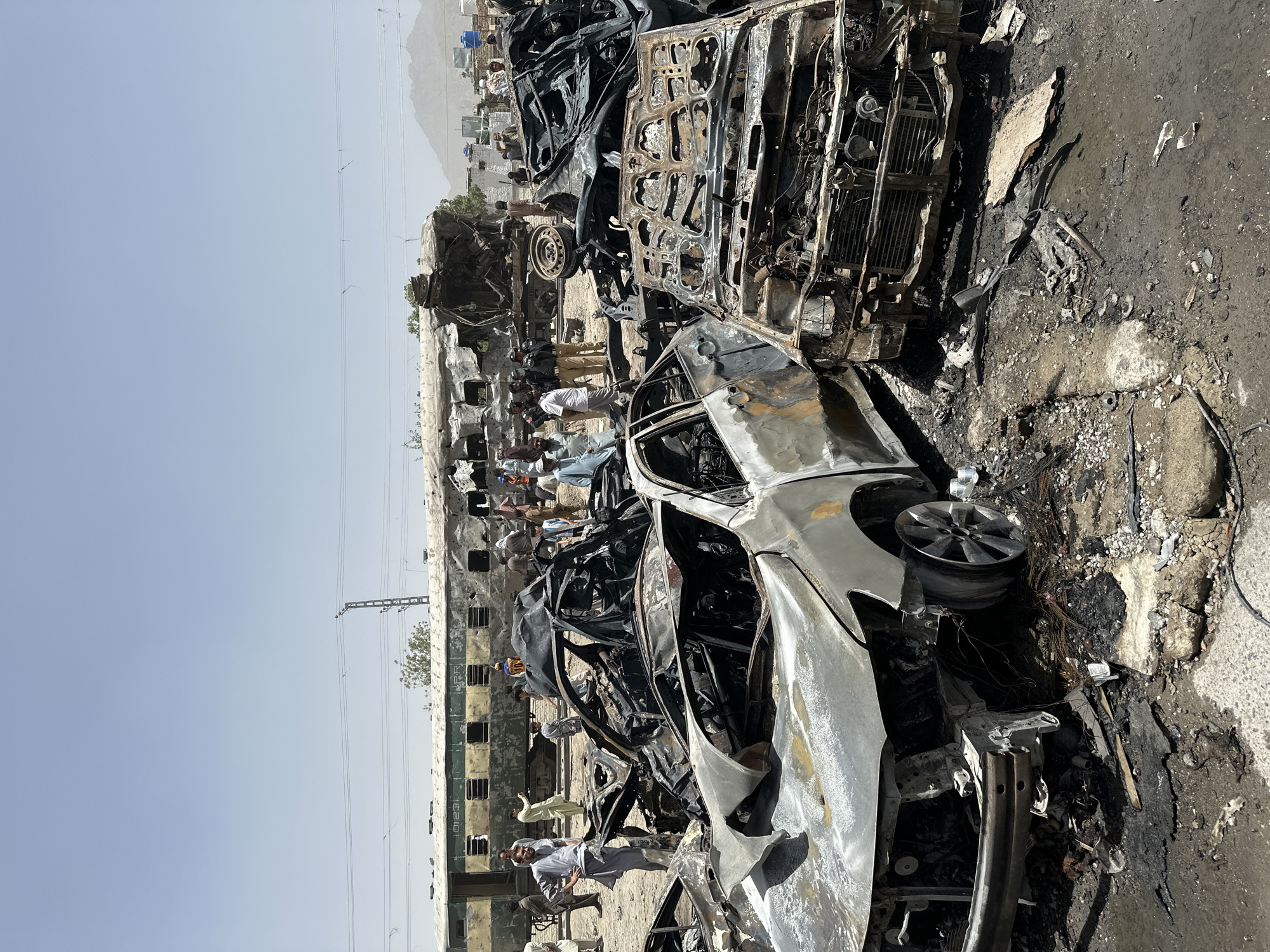 The image depicts a scene of destruction in Quetta City Tehsil, Pakistan. Several vehicles are heavily damaged, appearing burnt and partially disassembled. A group of individuals are standing nearby, observing the scene. In the background, buildings and mountains are visible under a clear sky. Overhead power lines stretch across the landscape. The environment seems arid, with dusty ground and debris scattered around the vehicles. A train is visible on the left side of the image. The overall scene suggests an aftermath of a violent event, likely involving fire and significant damage to property.
