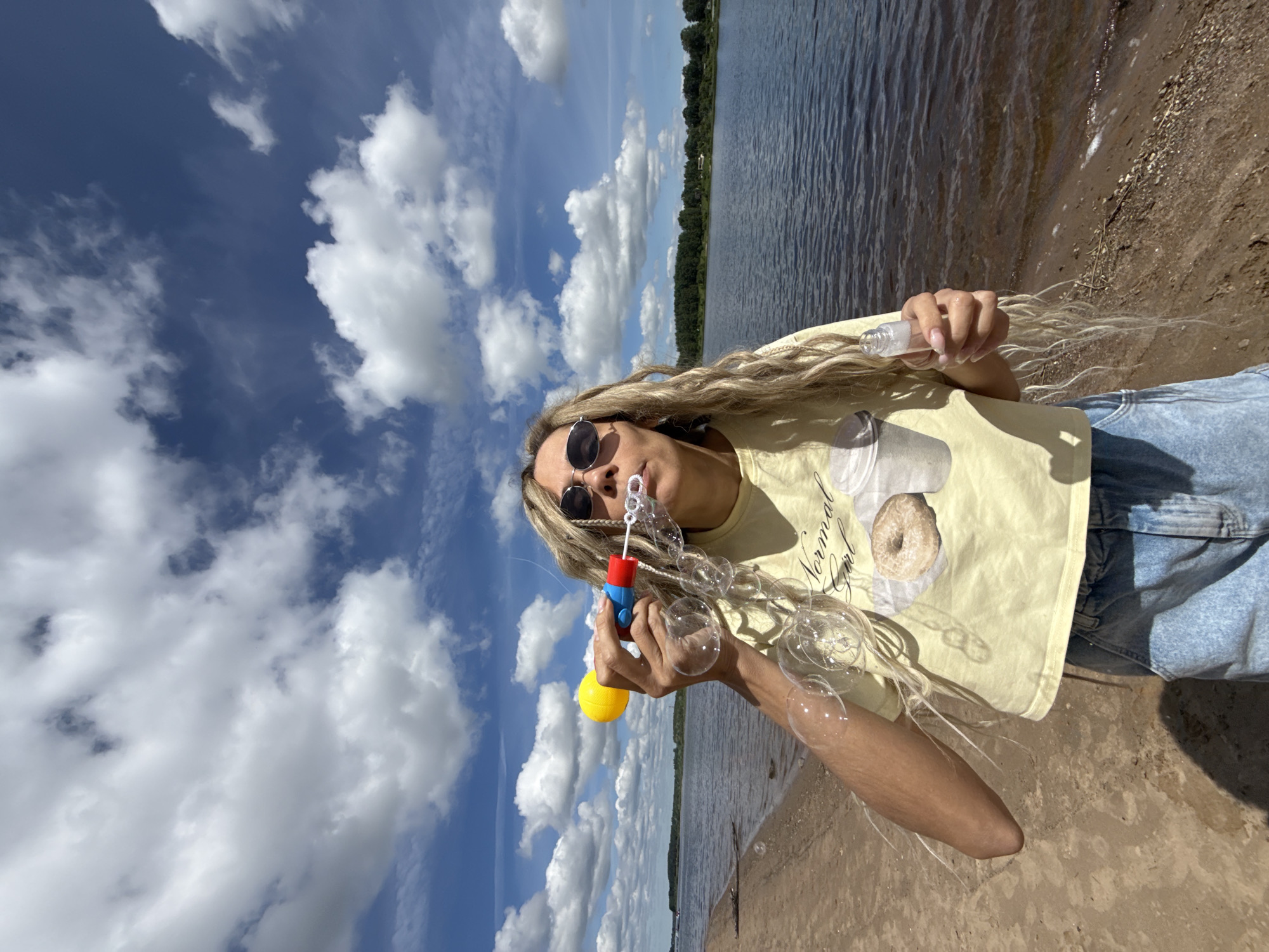A brightly lit outdoor scene, originally rotated 90 degrees clockwise, features a person with long, light-colored dreadlocks or wavy hair, actively blowing bubbles by a body of water in Rusanovo, Russia. The person wears dark sunglasses, a light yellow t-shirt, and blue denim jeans. In their left hand, they hold a colorful blue, red, and yellow bubble-making toy, from which numerous iridescent bubbles are emerging and floating. Their right hand holds a small, clear cylindrical bottle, likely containing bubble solution. On the yellow t-shirt, a graphic resembling a donut is visible, alongside the text "Normal Girl."
The setting is a sunny day under a bright blue sky filled with scattered, fluffy white cumulus clouds. To the person's right is a calm body of water, appearing to be a lake or wide river, with gentle ripples on its dark surface. A sandy or earthy shoreline, light brown in color, forms the immediate ground. Across the water in the background, a line of lush green trees marks the distant bank. The overall atmosphere is one of leisure and playfulness on a clear, pleasant day.