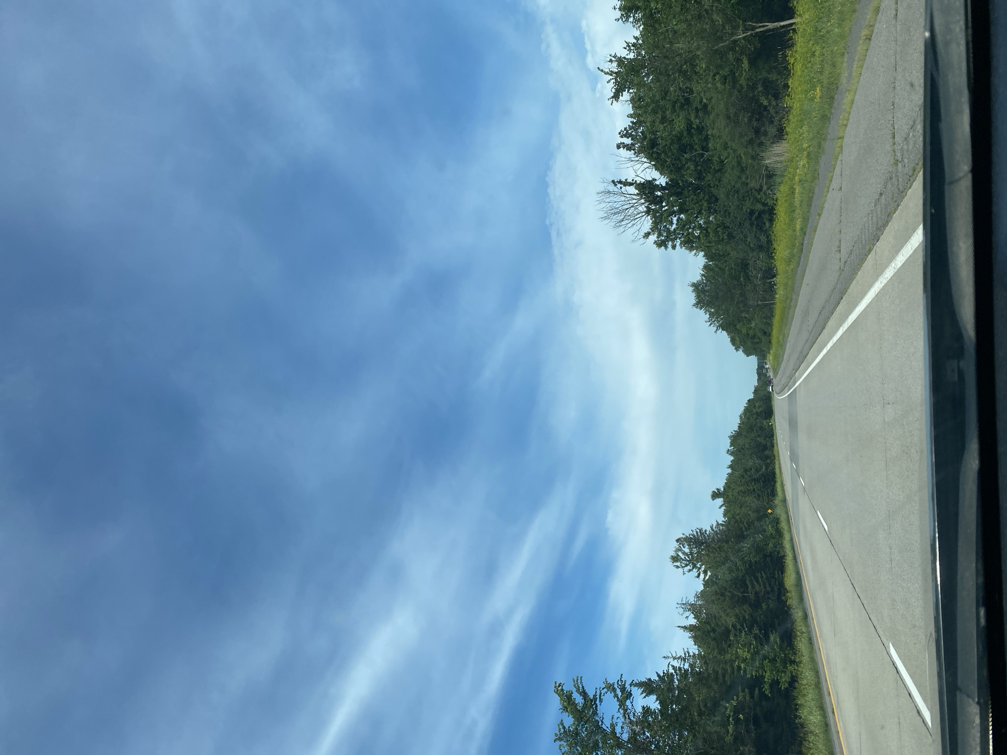 The image shows a view from the perspective of a vehicle traveling on a highway. The highway is paved and has lane markings, including a yellow line on the left and white lines separating the lanes. On either side of the highway, there are dense green trees and some yellow wildflowers. The sky is a bright blue with thin, wispy white clouds. The overall environment appears to be a rural or semi-rural setting. There are no people or other vehicles clearly visible in the immediate foreground or midground, though distant vehicles can be seen on the highway.