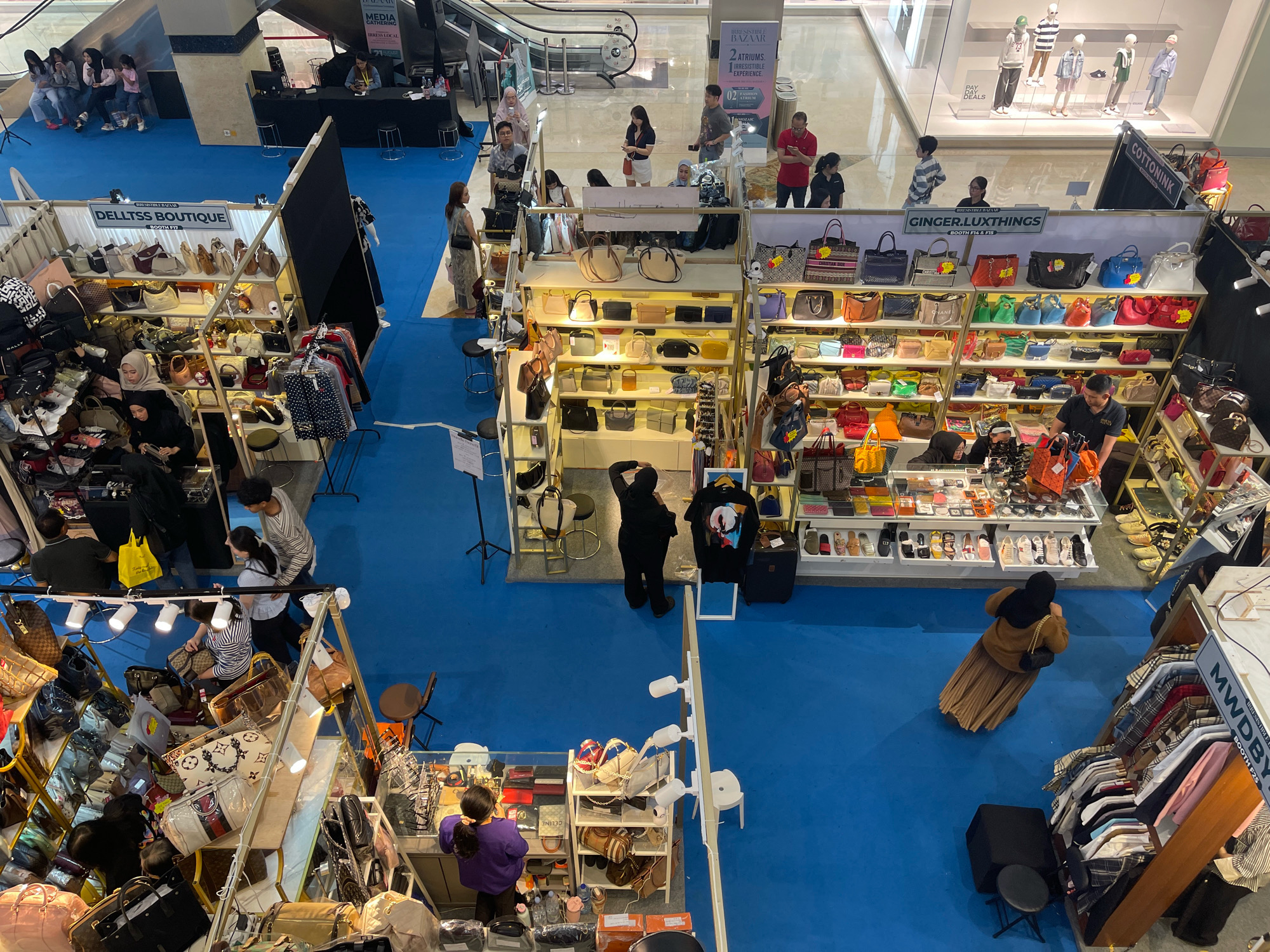 An aerial view captures a bustling scene on a blue carpeted floor inside a modern indoor shopping mall, possibly at Kota Kasablanka in South Jakarta, Indonesia. The space is filled with numerous retail booths showcasing a variety of goods, primarily bags and clothing. Several people are present, some browsing merchandise, others manning the stalls, and a few are seen walking through the aisles.
In the foreground, to the left, is a stall overflowing with handbags and accessories. A person in a dark hijab is seated behind the counter, interacting with a customer. Further back, a man in a black shirt and a woman in a brown hijab are engaged in conversation while standing near a display of bags.
To the right, a row of clothing racks displays various patterned shirts, with a woman in a brown hijab walking away from the camera. Above this area, another stall, "GINGER.LUXTHINGS," is visible, featuring rows of colorful handbags.
In the background, a large escalator is present, indicating a multi-level mall. More retail booths and individuals can be observed in the distance, contributing to the lively atmosphere of the marketplace. The overall scene depicts a busy shopping event or market taking place within a commercial establishment.